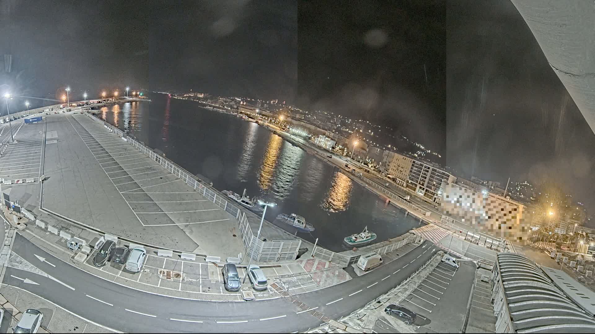 A nighttime aerial view of a harbor with a mostly empty parking lot, several small boats on the water, and city lights in the background.