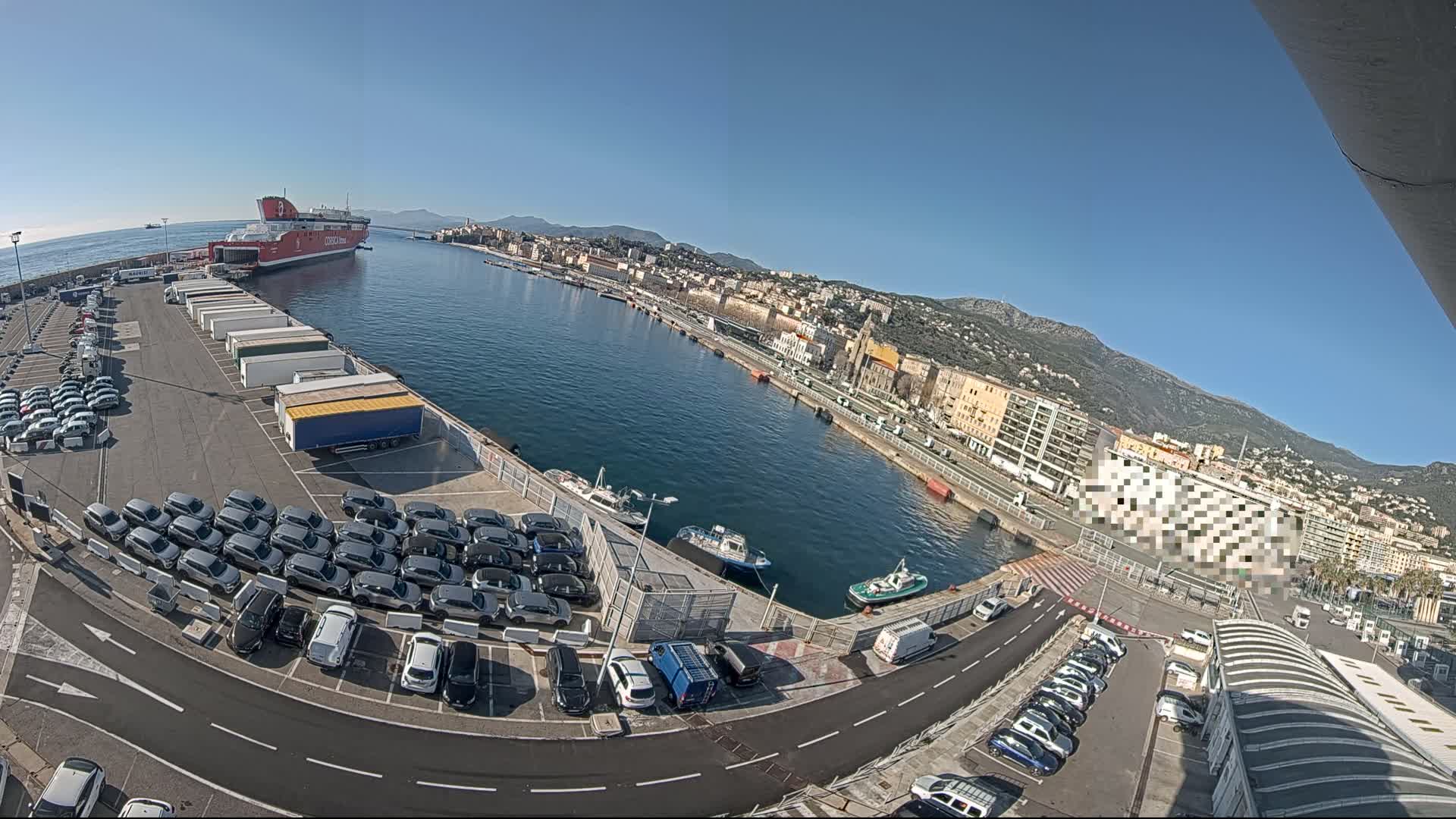 A large red ferry is docked at a port city on a sunny day, with numerous cars parked in lots surrounding the harbor, and smaller boats in the water.
