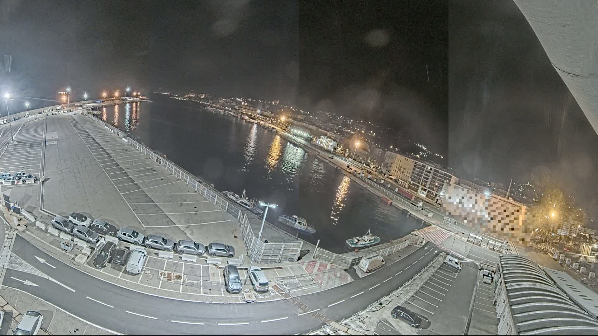 A nighttime aerial view of a harbor with a parking lot full of cars, several small boats on the water, and city lights in the distance.