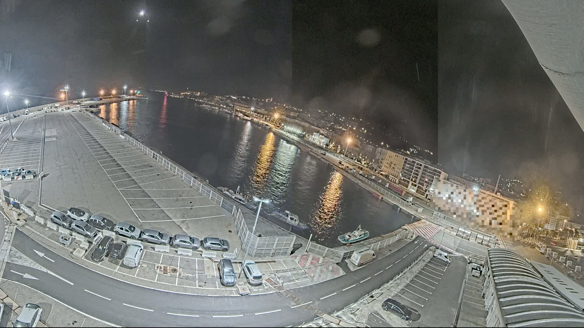A nighttime aerial view of a harbor with a mostly empty parking lot, several small boats on the water, and city lights in the background under a clear, moonlit sky.
