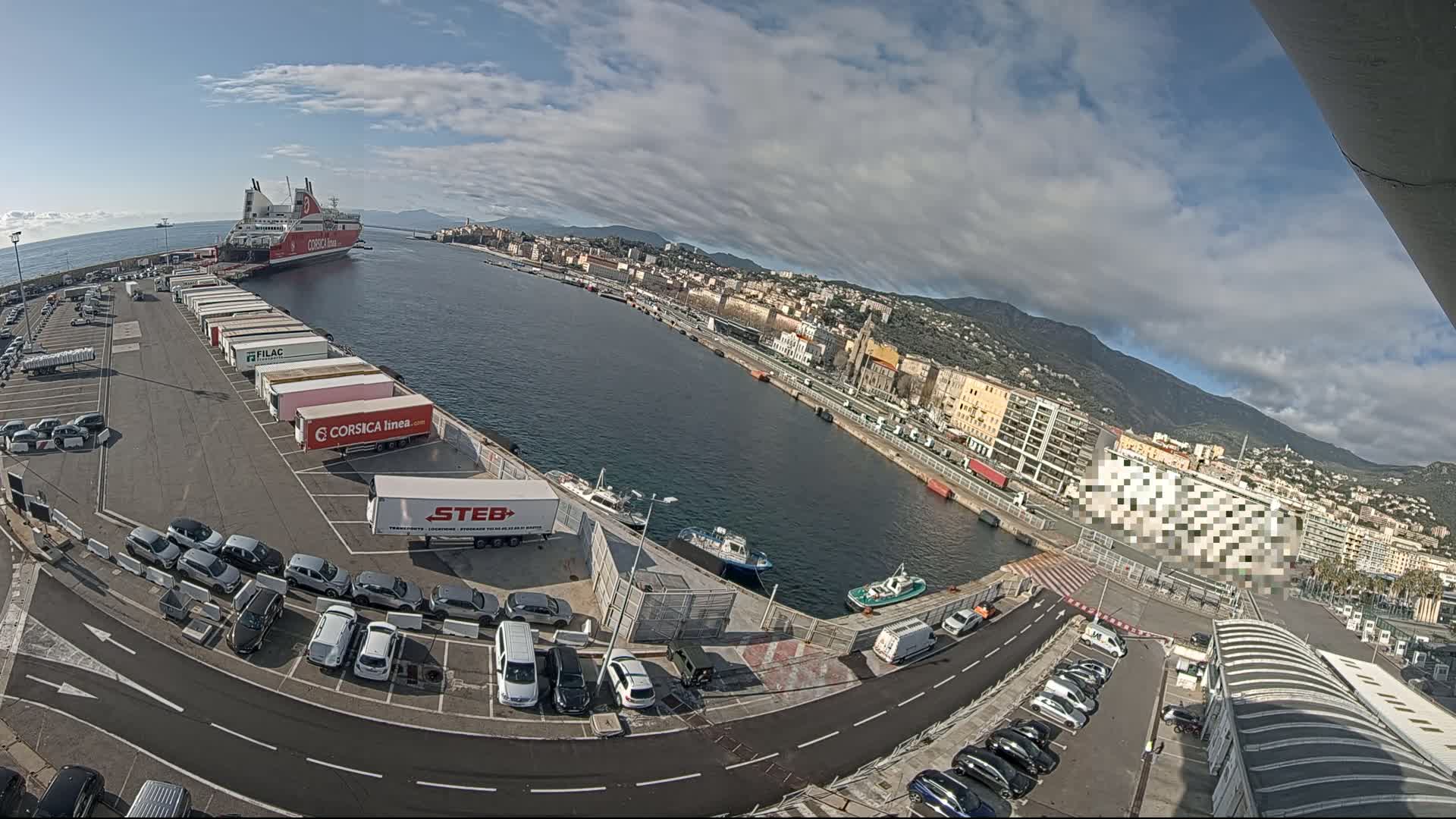 A ferry is docked at a port on a partly cloudy day, with numerous cars and trucks parked nearby, and a city visible in the background.