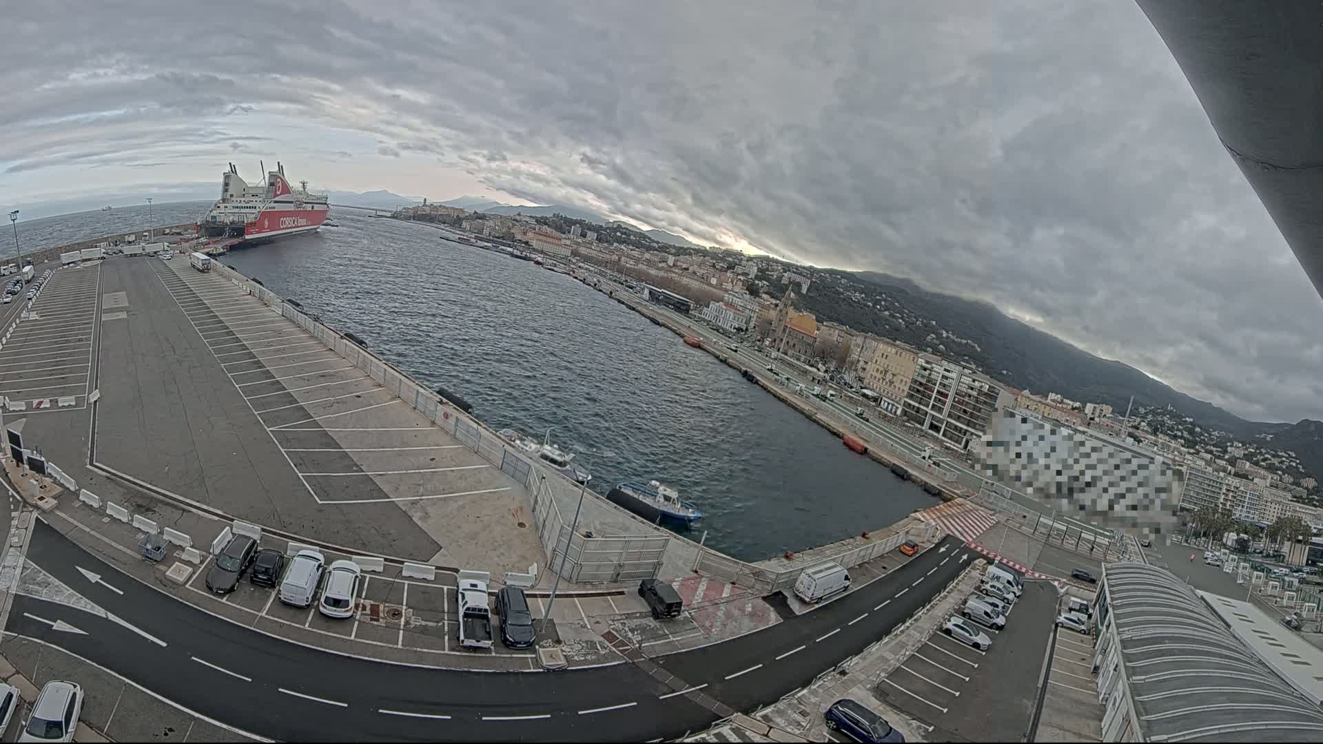 A large ferry is docked at a port, with overcast skies and a city visible in the background.