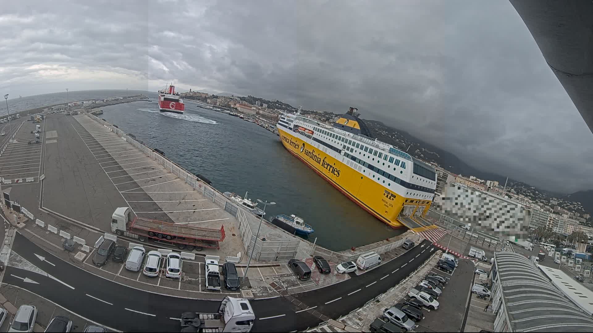 A large yellow ferry is docked at a port with several cars parked nearby, under an overcast sky.