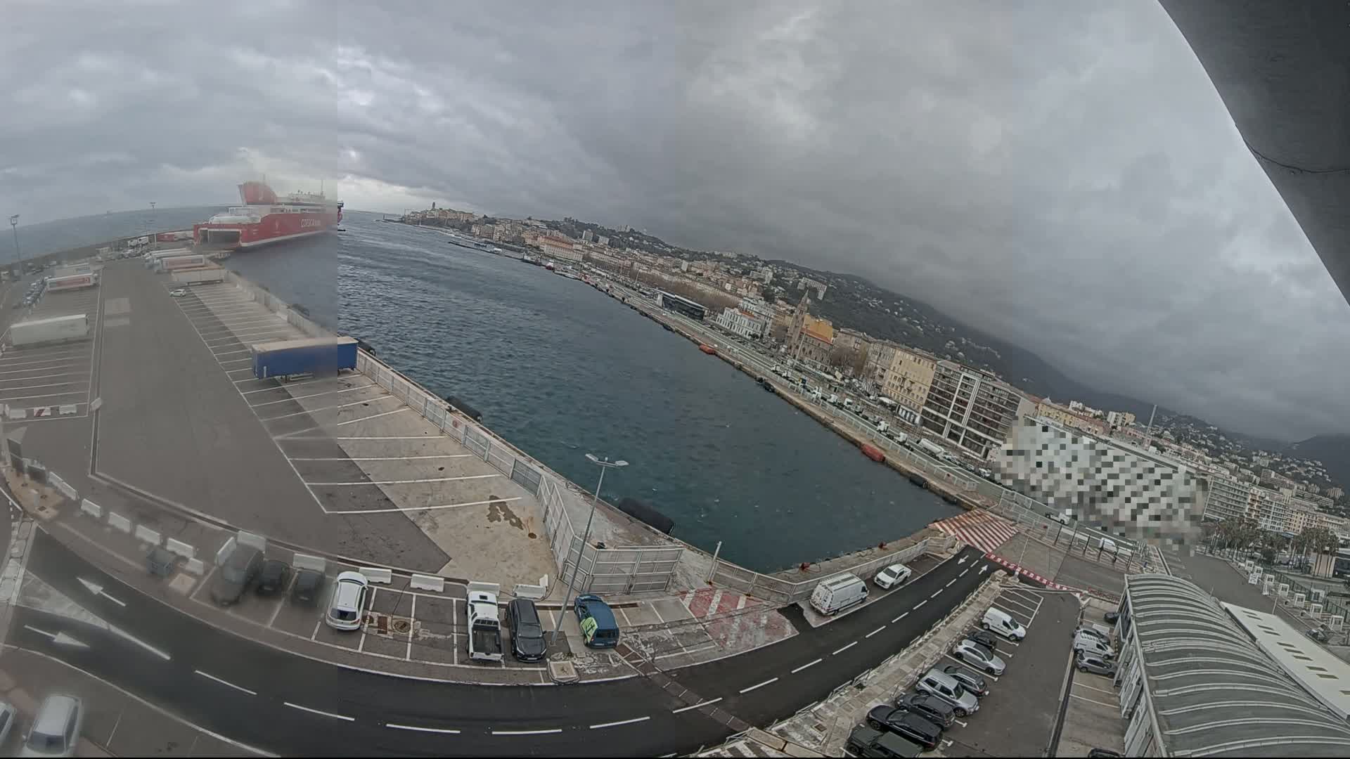 A large red ferry is docked at a port city under a cloudy sky, with cars parked in lots surrounding the harbor.