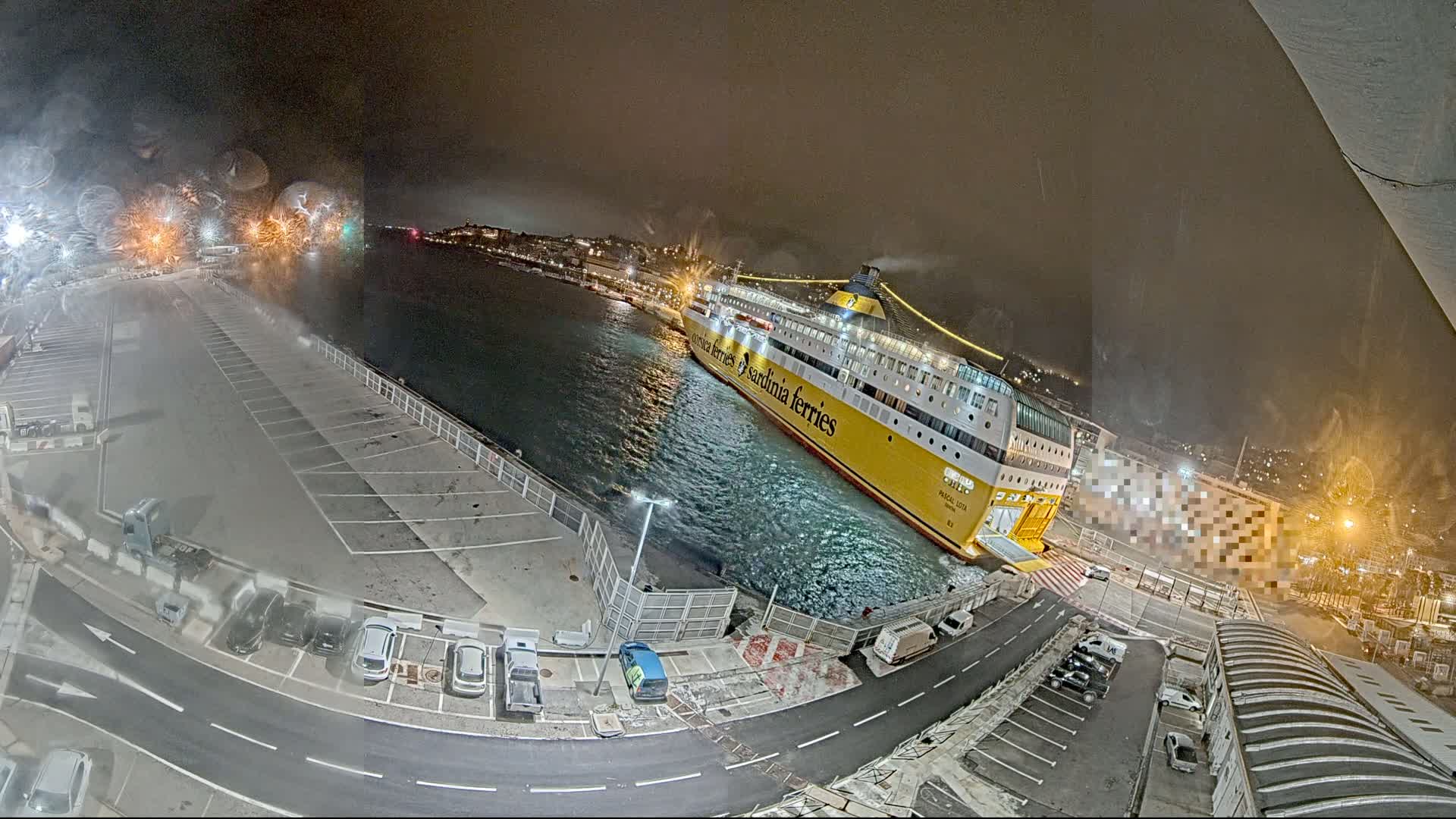 A large yellow and white ferry is docked at night in choppy water, near a parking lot and other buildings.