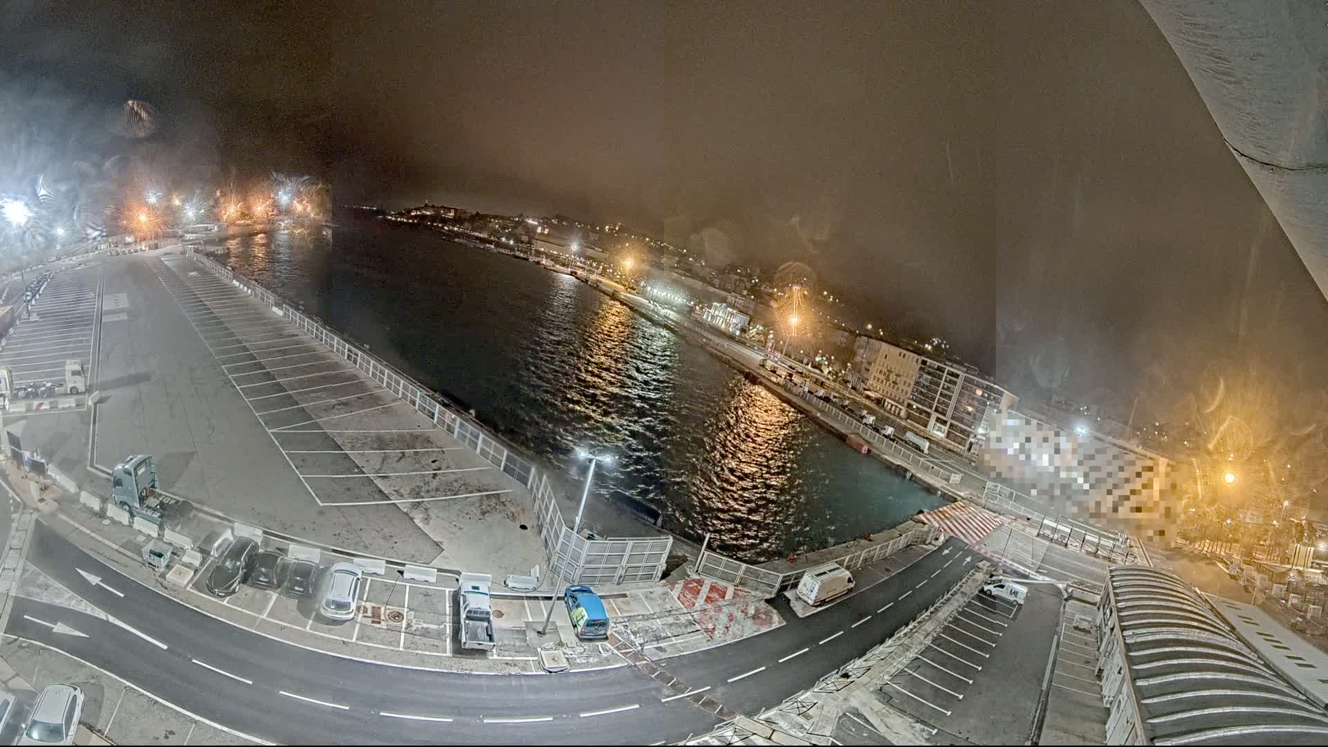 A nighttime, aerial view of a harbor with a mostly empty parking lot, a roadway, and a body of water reflecting city lights under a hazy sky.
