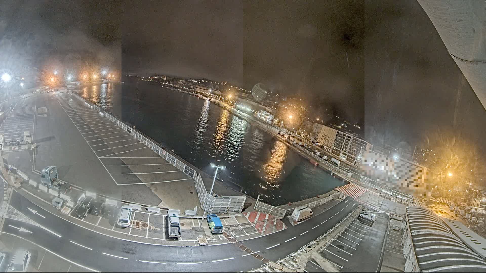 A nighttime aerial view of a harbor with a mostly empty parking lot, several vehicles, and a body of water reflecting city lights under hazy conditions.