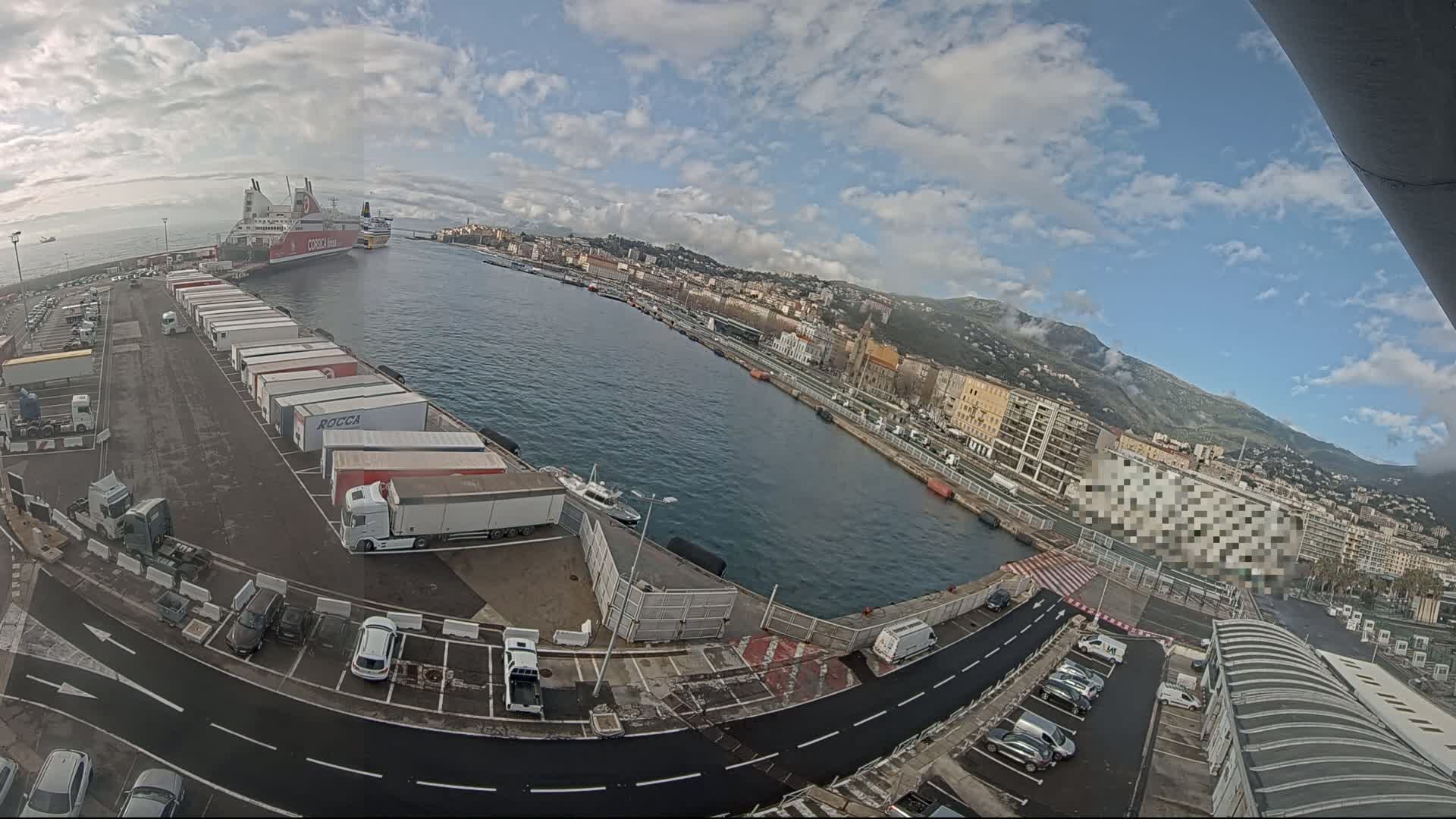 A partly cloudy day reveals a harbor scene with a large ferry docked near numerous semi-trucks and cars parked in a lot, all overlooking a city nestled against a mountain range.