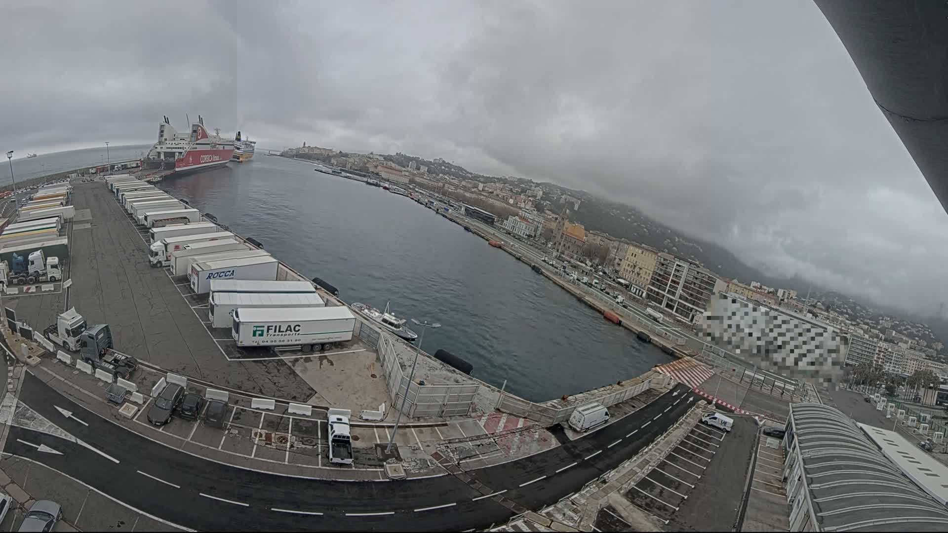 A ferry is docked at a port city on an overcast day, with numerous semi-trailer trucks parked nearby.