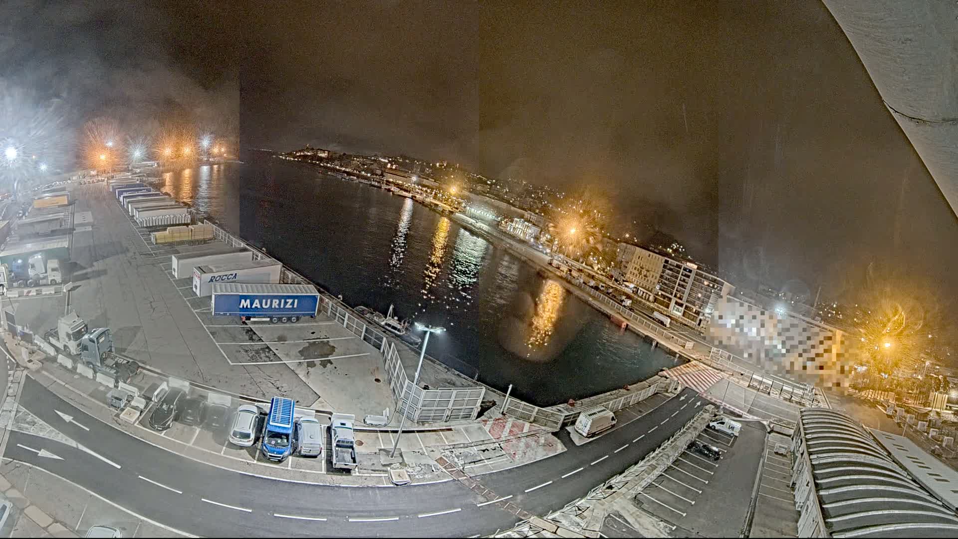 A nighttime, high-angle view of a harbor with trucks, cars, and a body of water reflecting city lights under a hazy sky.