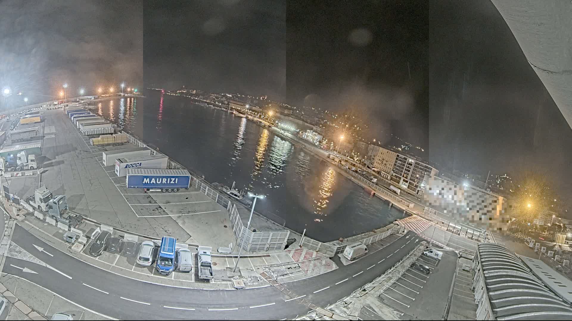 A nighttime aerial view of a harbor shows a calm body of water reflecting city lights, with parked trucks and cars visible on the dock.