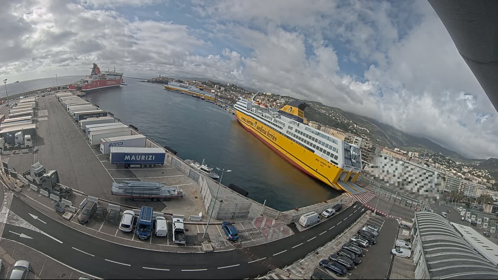 A large yellow ferry is docked at a port, with several other ships and numerous trucks and cars in the parking lot under a partly cloudy sky.