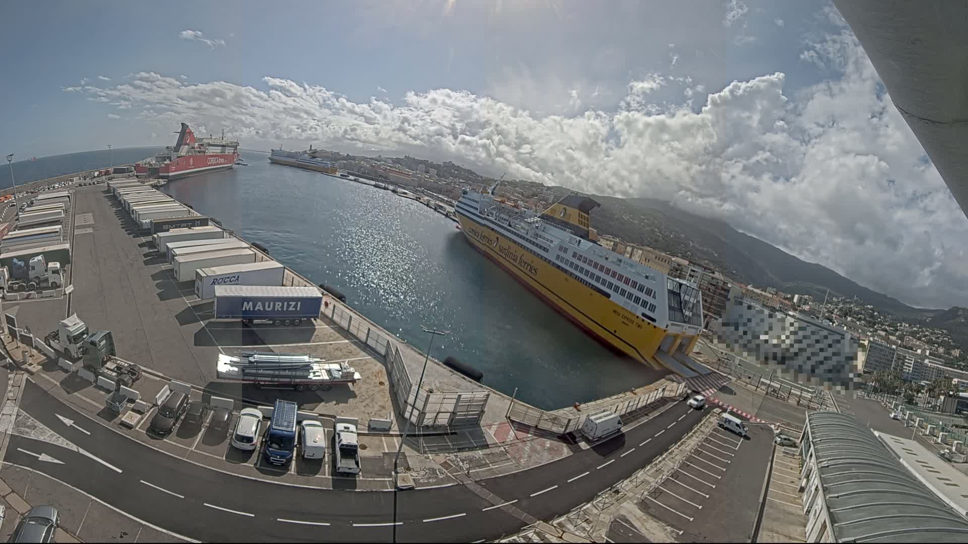 A mostly sunny day at a port shows two large ferries docked, one red and one yellow, with several trucks and cars parked nearby.