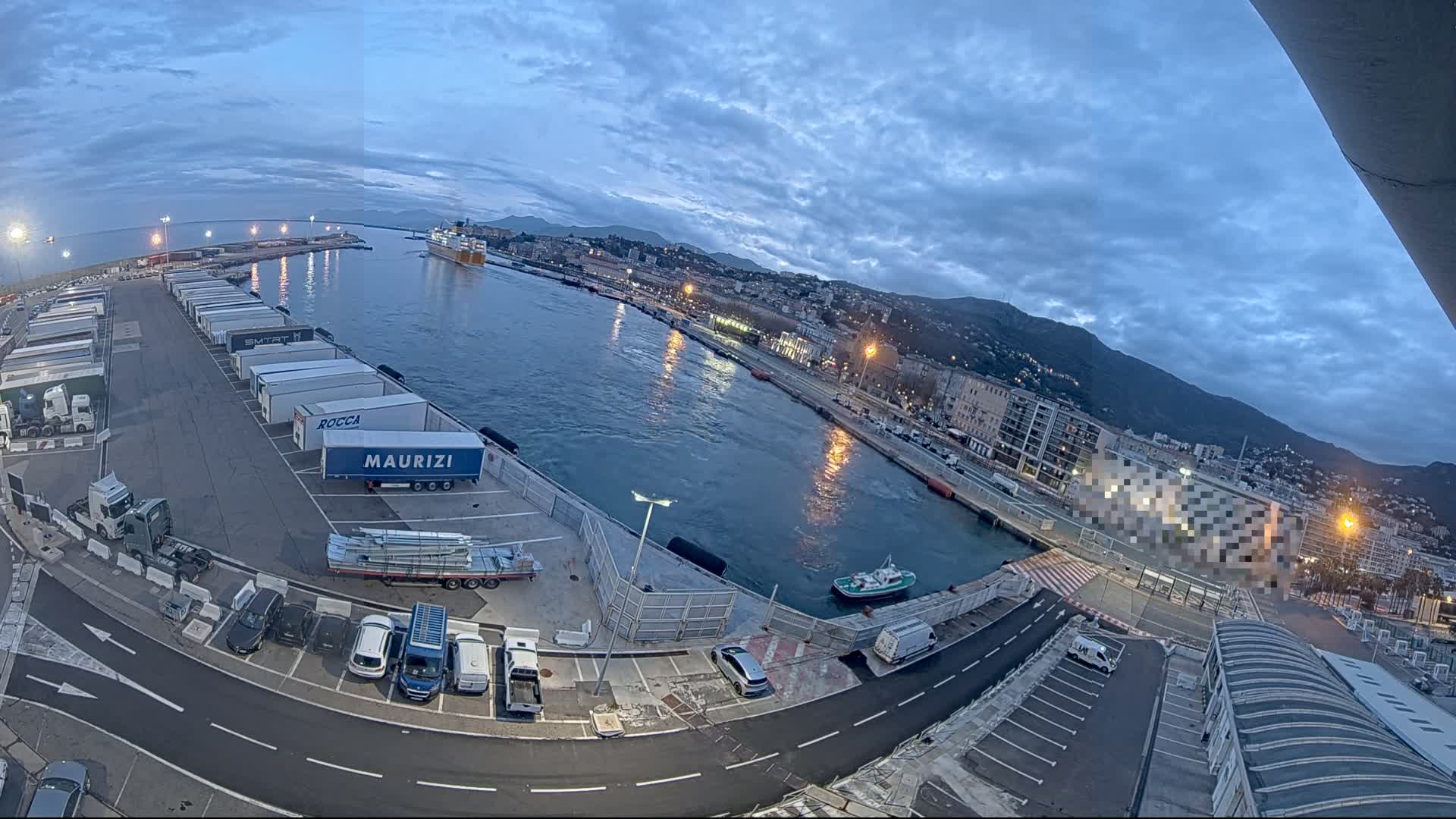 A harbor at dusk, with a large cargo ship in the distance, numerous semi-trailer trucks in a parking lot, several cars, and a small harbor boat in the water.