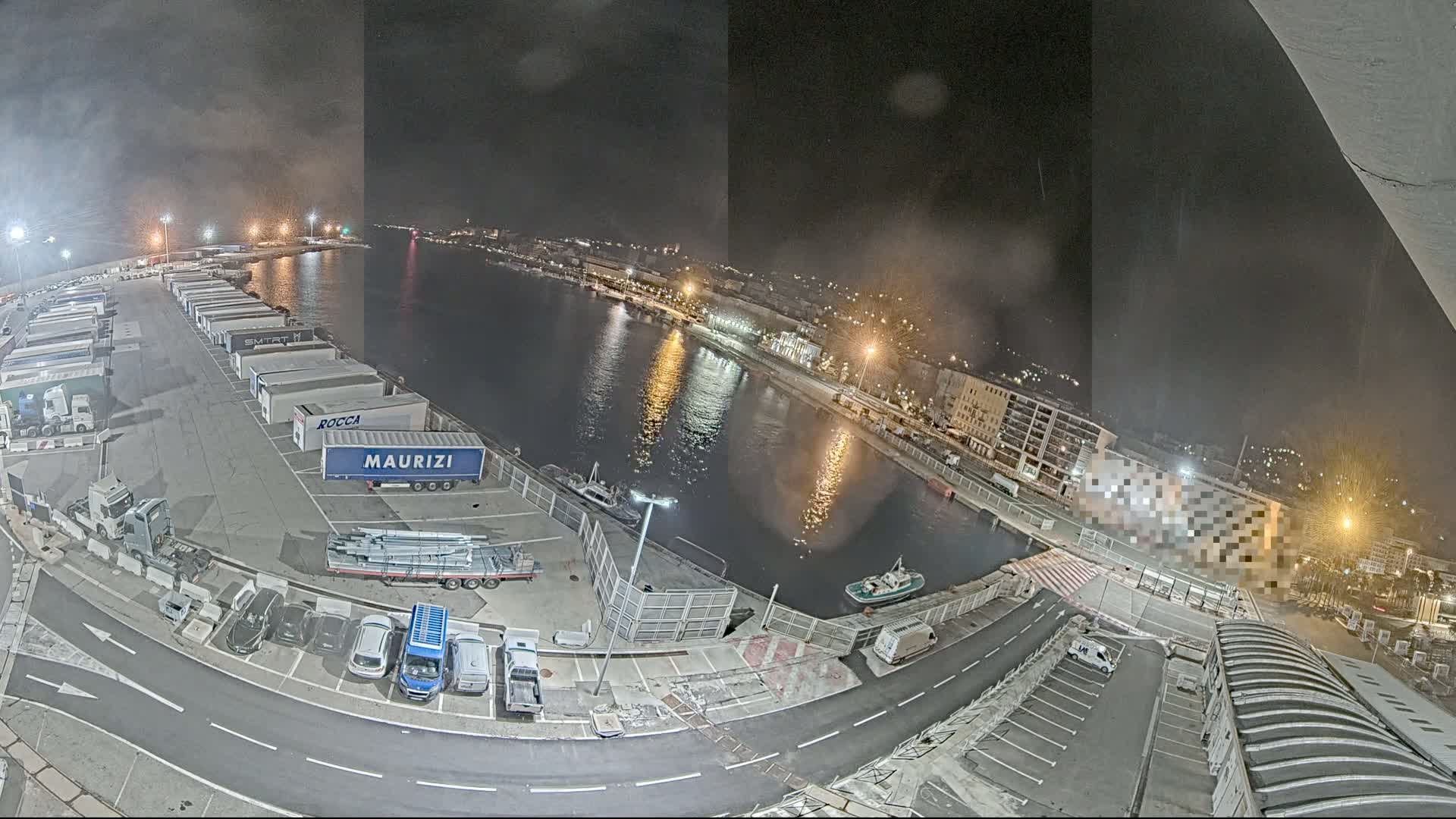 A nighttime aerial view of a harbor shows a parking lot filled with semi-trailer trucks, cars, and a calm body of water reflecting city lights.