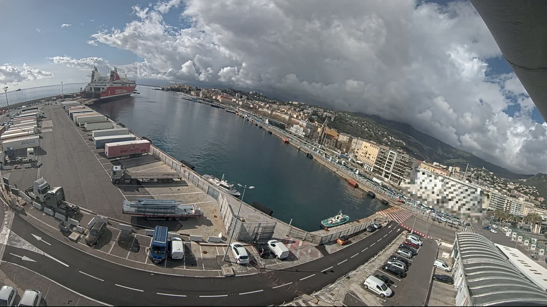 A partly cloudy day shows a harbor with a large ferry docked, numerous trucks and cars parked in a lot, and a city stretching along the waterfront under a partly cloudy sky.