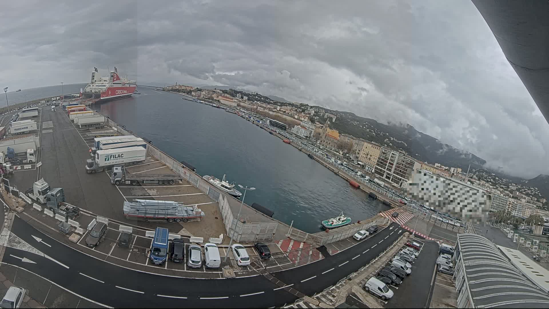 A ferry is docked at a harbor with overcast skies, surrounded by numerous trucks, cars, and smaller boats, all situated against a backdrop of a city and mountains.