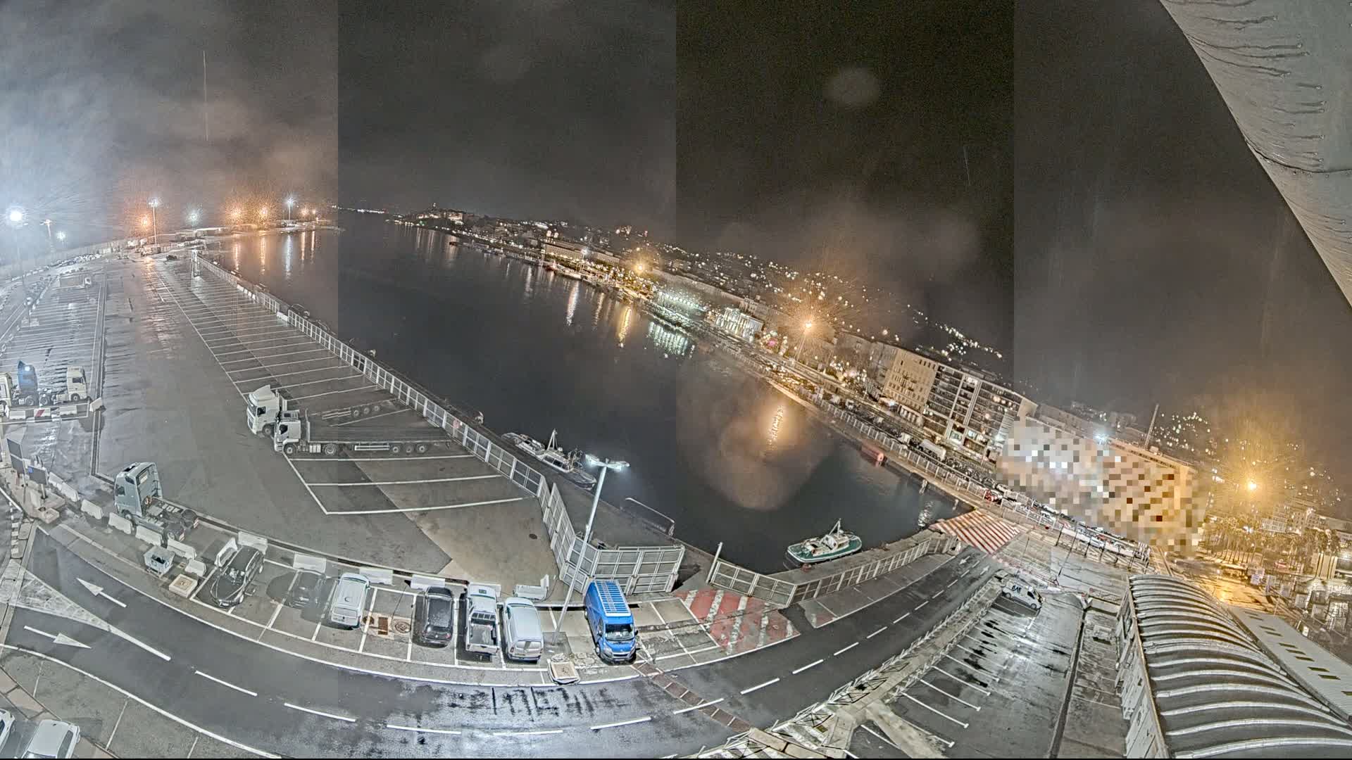 A nighttime view of a harbor with a wet parking lot containing several vehicles, boats in the water, and a city skyline in the background under foggy conditions.