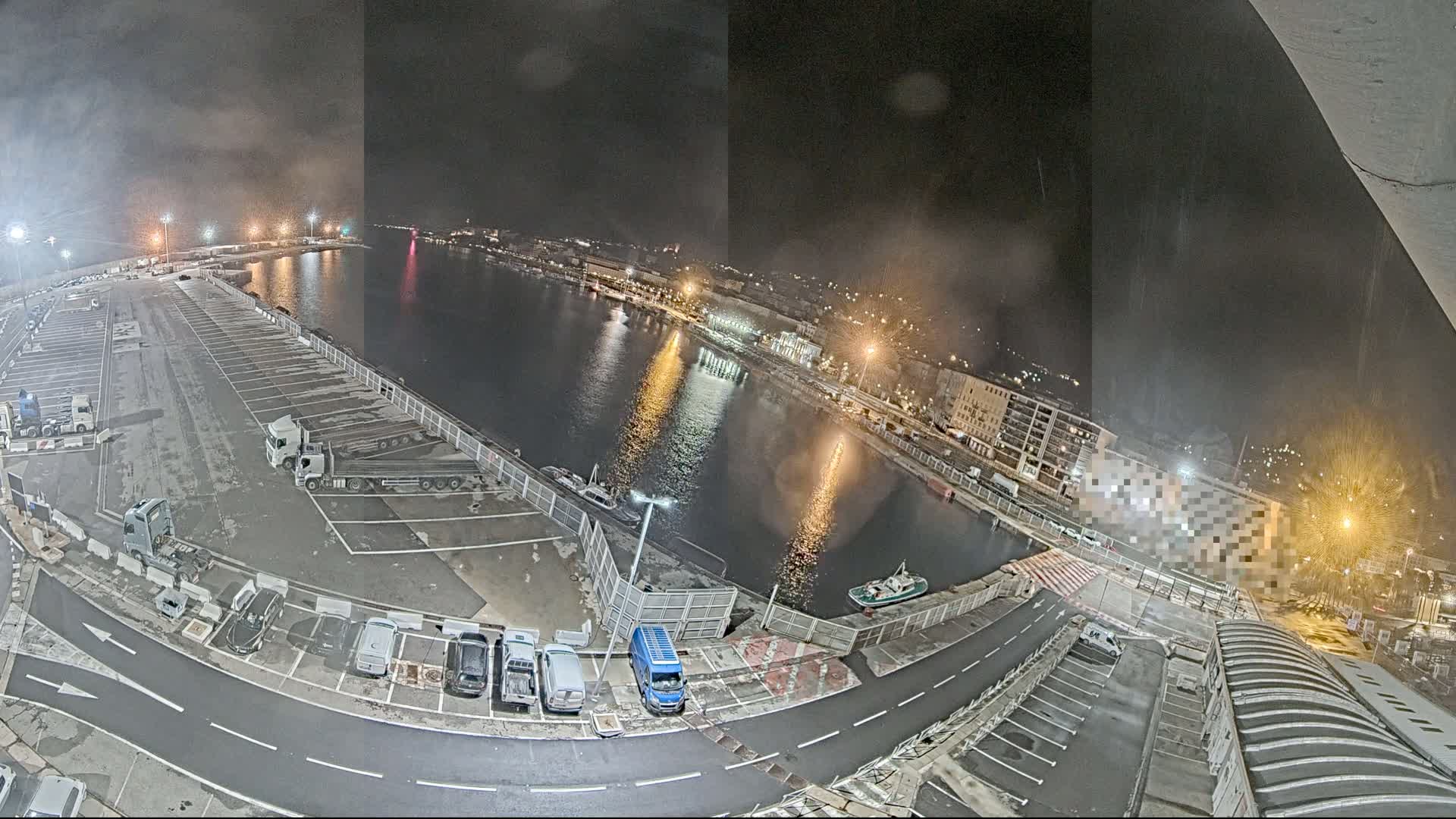 A nighttime aerial view of a harbor with a mostly empty parking lot filled with a few cars and trucks, a calm body of water reflecting city lights, and buildings lining the waterfront.
