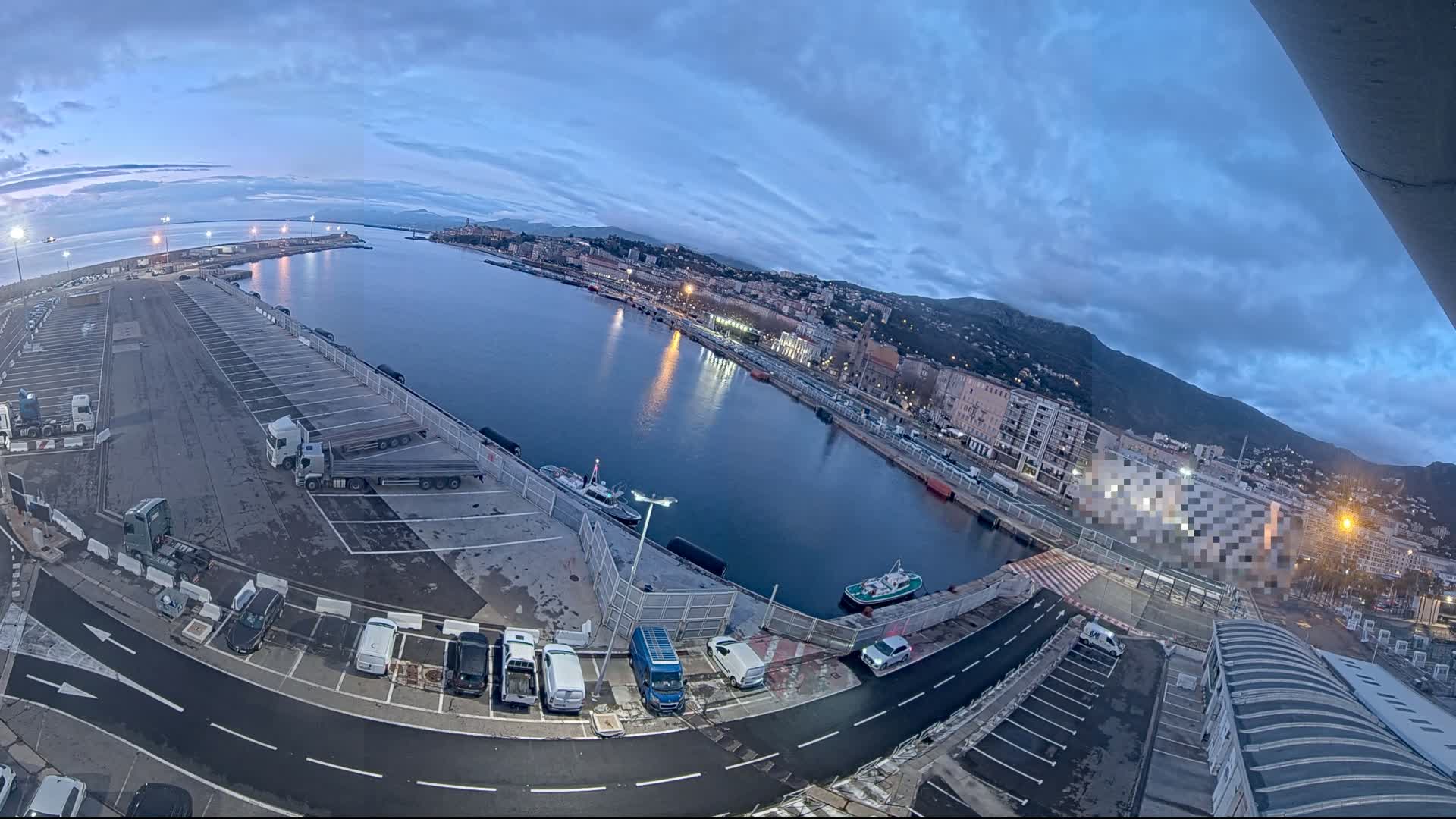 An aerial, wide-angle view of a harbor at dawn, showing a calm body of water reflecting the city lights, with parked vehicles and trucks in the foreground.