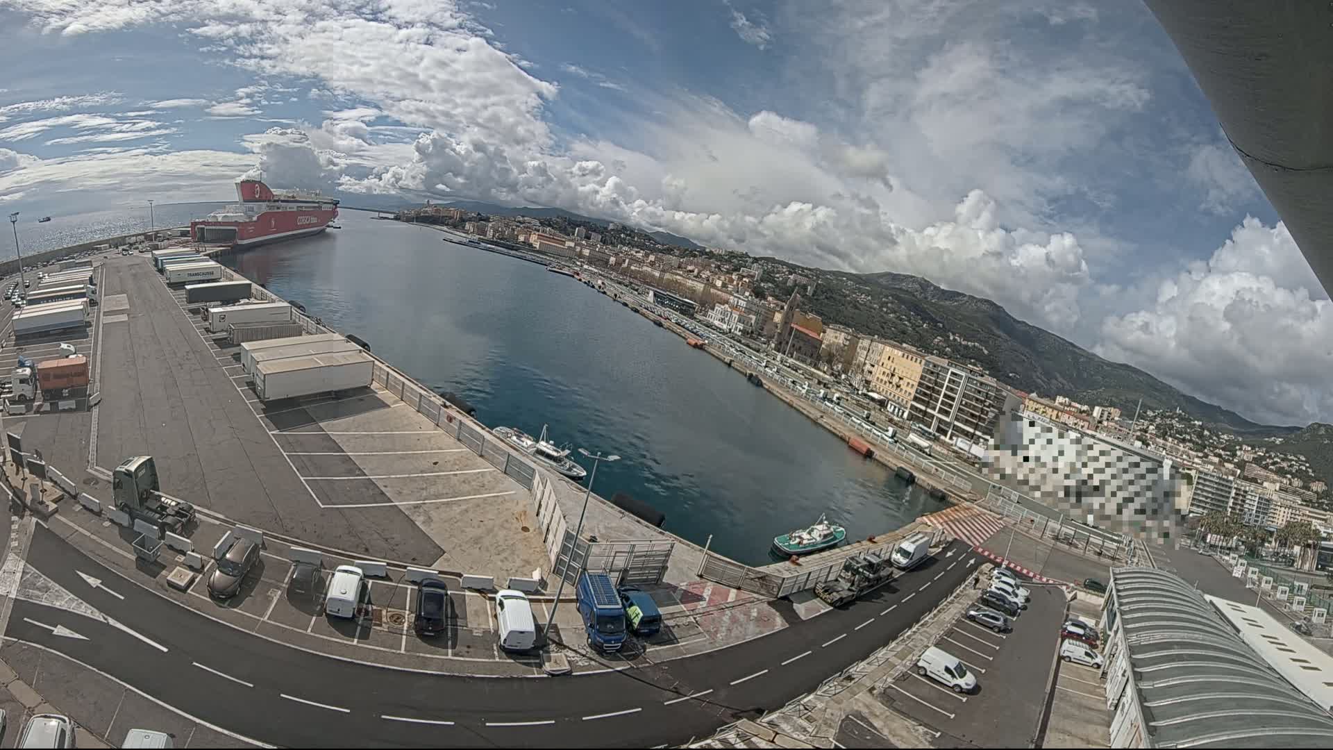 A partly cloudy day reveals a harbor with a large red ferry docked, numerous vehicles parked in lots, and a city sprawling on the hillsides in the background.