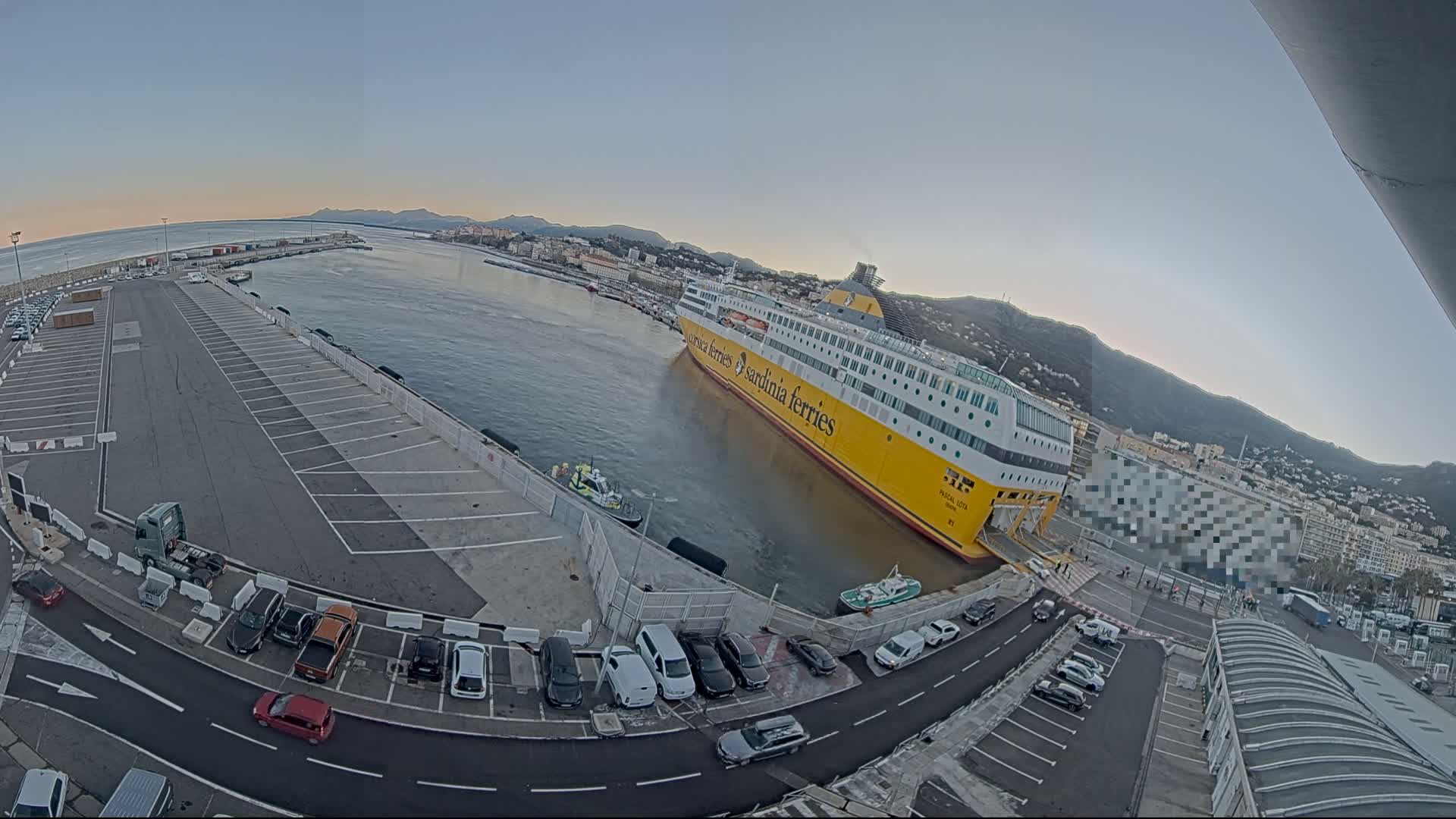A large yellow ferry is docked at a port, with cars parked in a lot nearby, under a clear, twilight sky.