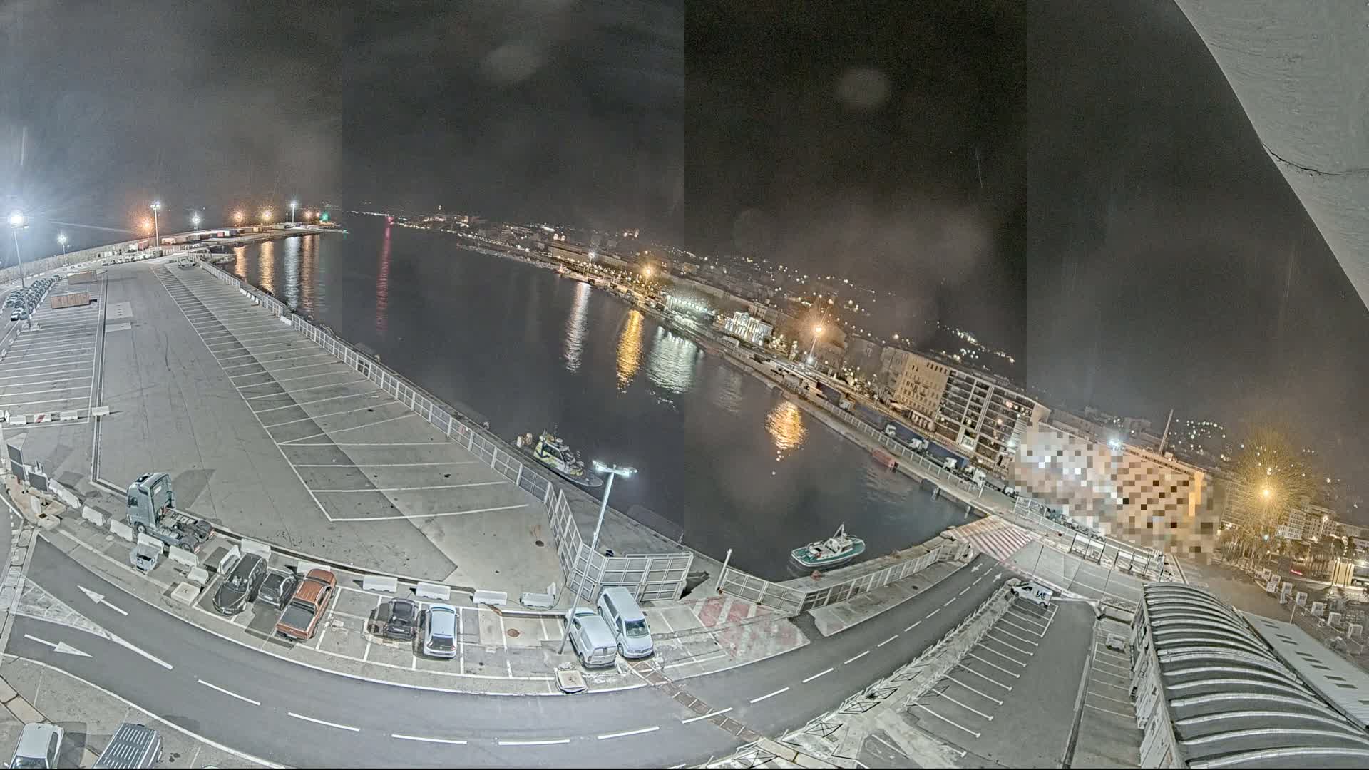 A nighttime aerial view of a harbor with a mostly empty parking lot, two small boats on the water, and city lights reflected in the calm water.