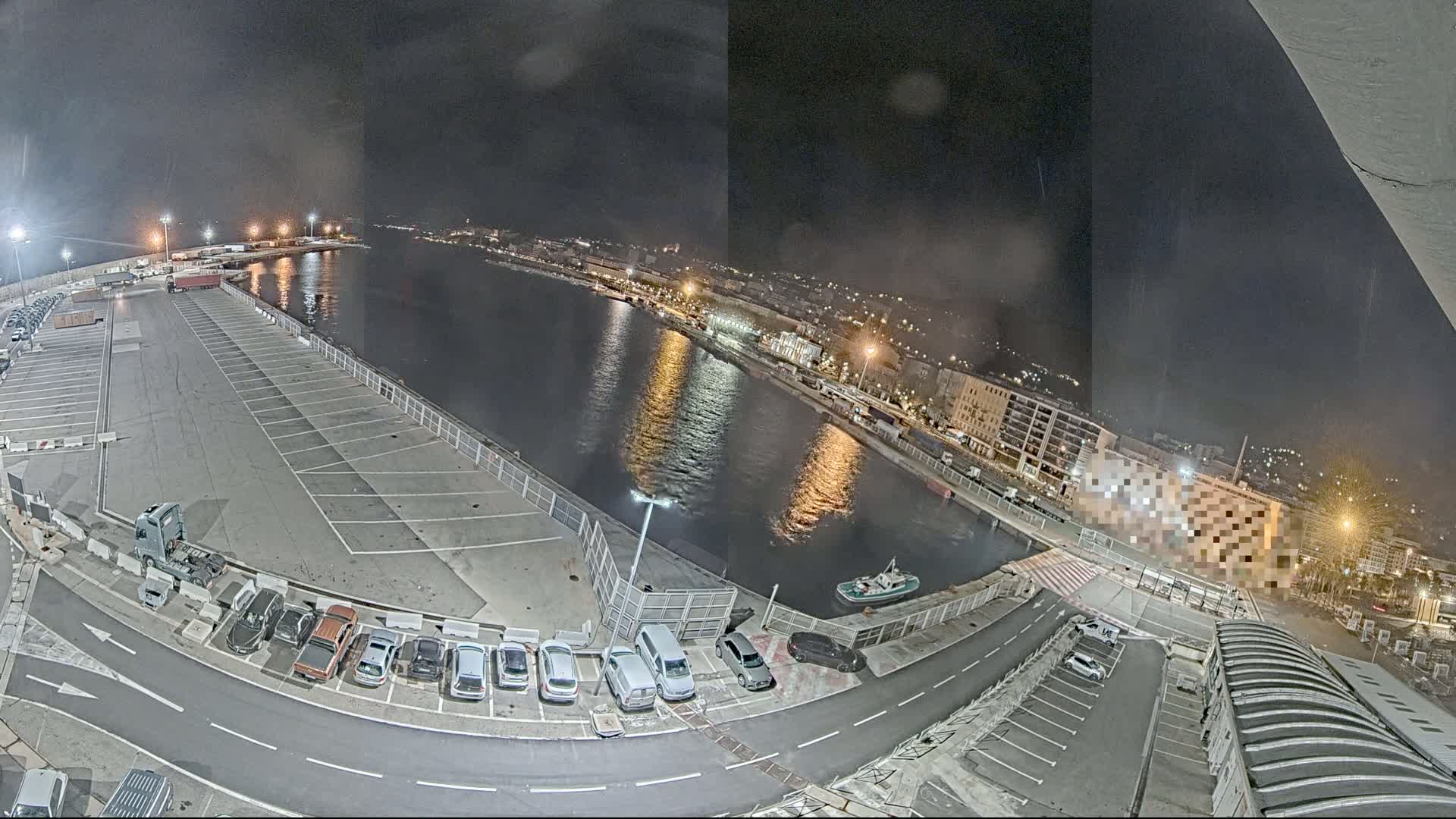 A nighttime aerial view of a harbor with a large empty parking lot, several cars parked along a roadway, and a small tugboat on the water.