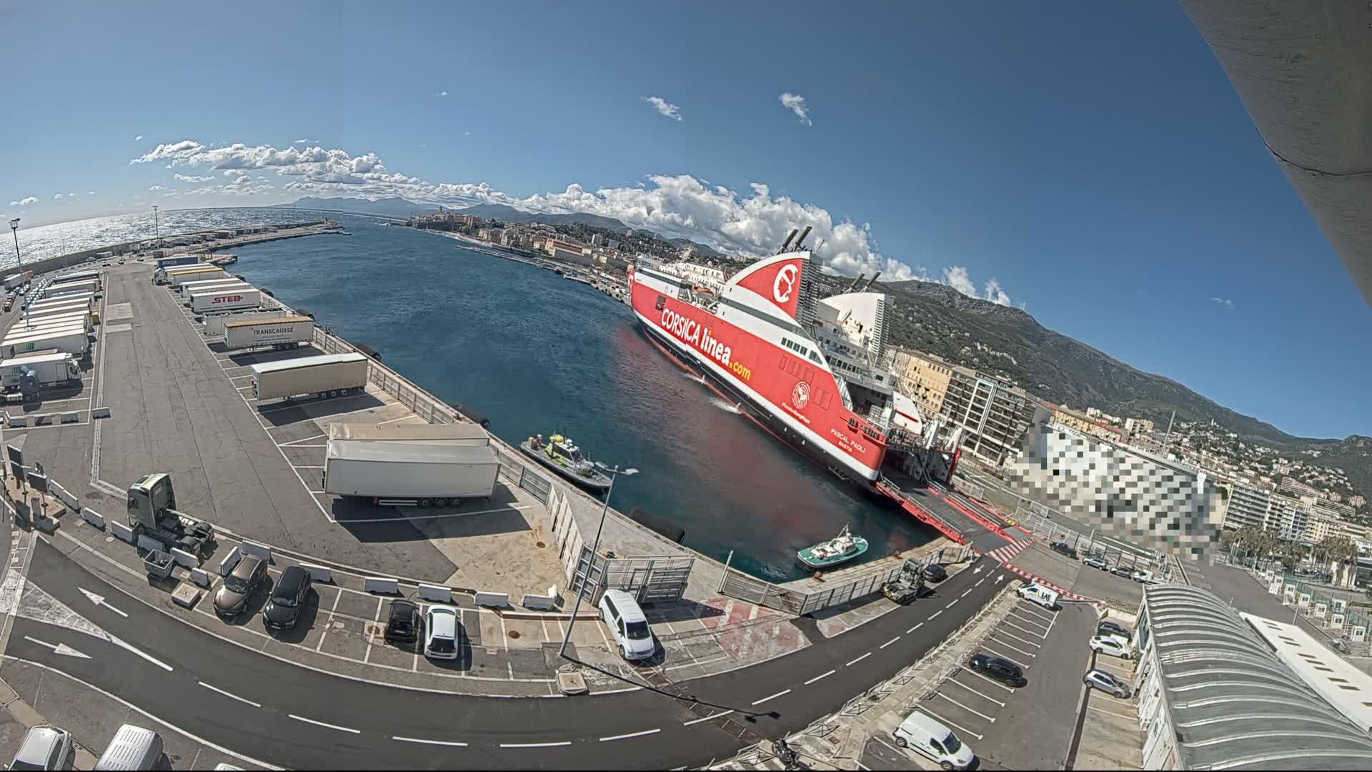 A large red and white ferry is docked at a port on a sunny day with partly cloudy skies, surrounded by parked cars and trucks.