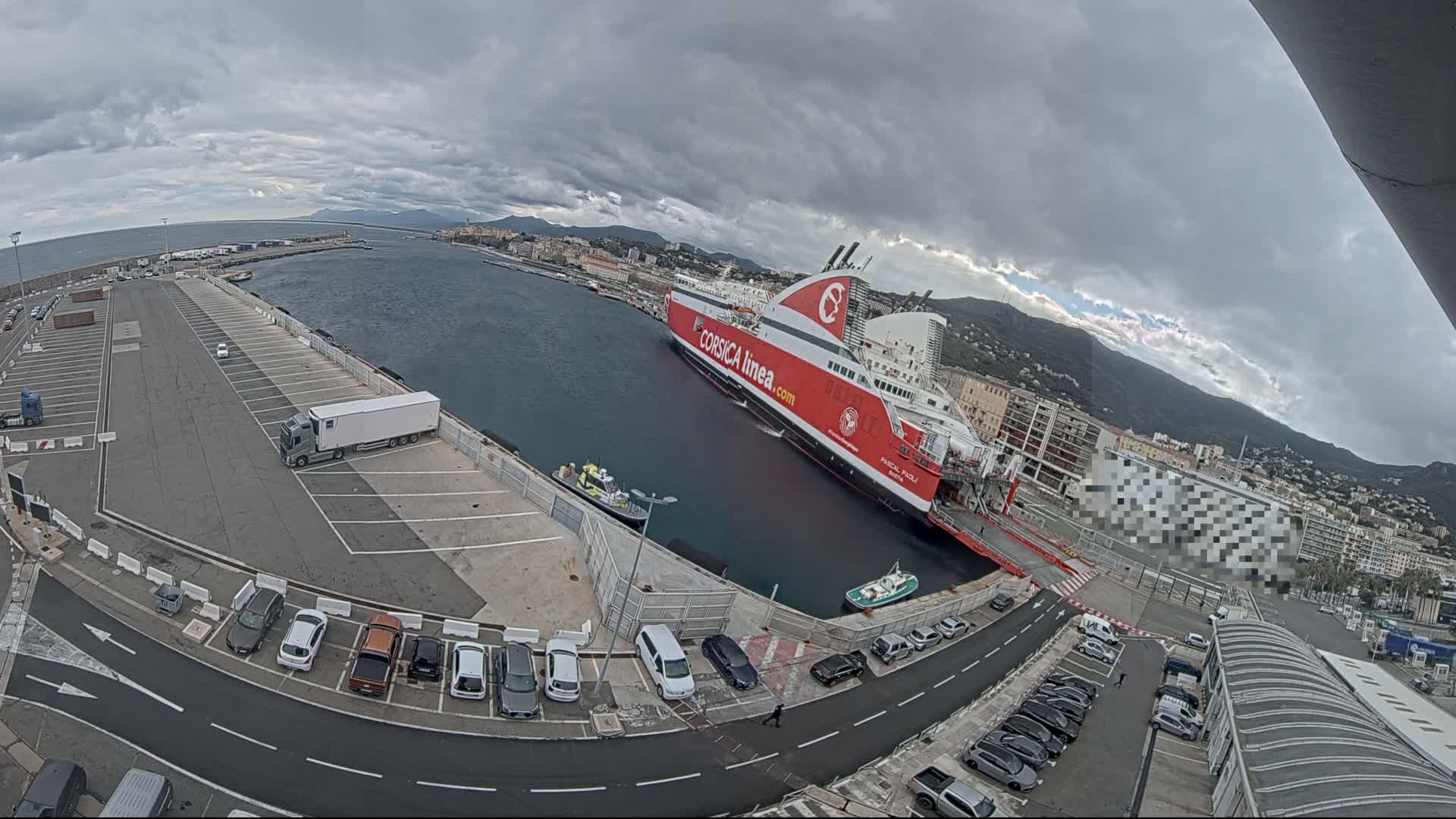 A large red and white ferry is docked at a port under a cloudy sky, with several cars parked in nearby lots and a truck in the parking area.