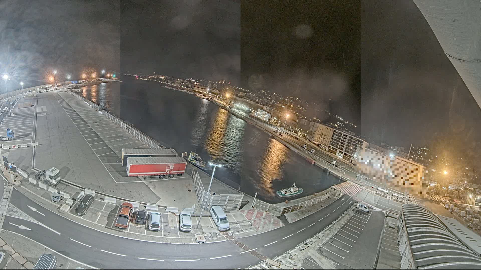 A nighttime aerial view of a harbor with a large empty parking lot, several vehicles, and a few small boats on the water.