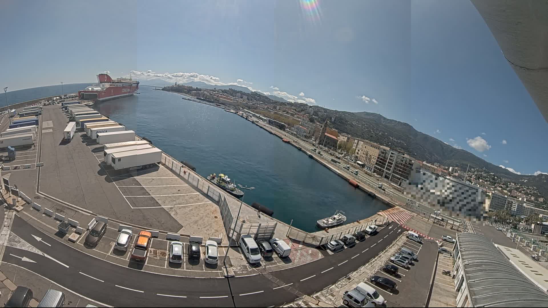 A large red ferry is docked at a harbor on a sunny day, with numerous cars and trucks parked in the surrounding lots, and small boats in the water.