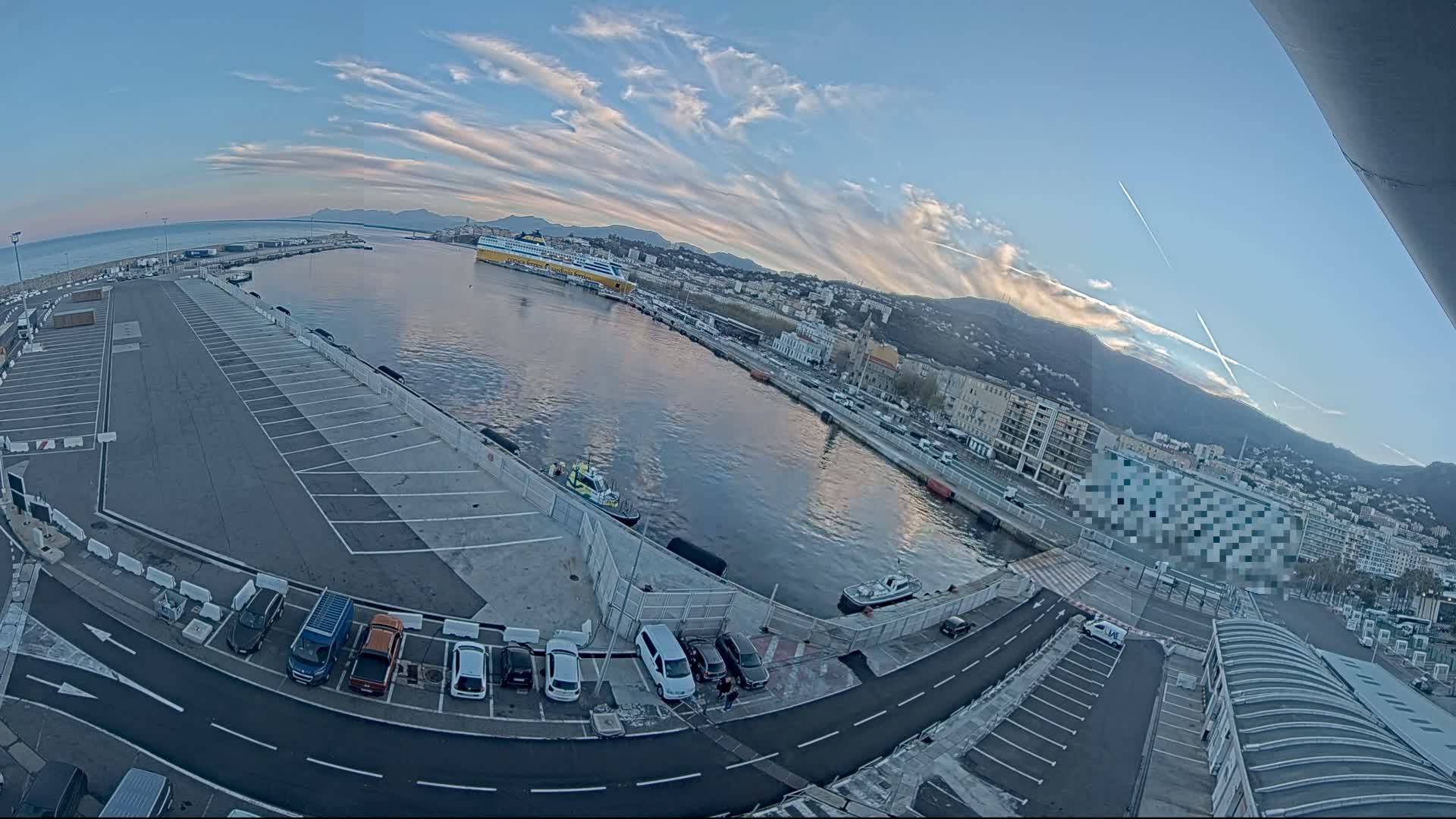 A harbor with a large parking lot, a ferry, and several small boats under a mostly clear sky with thin clouds.