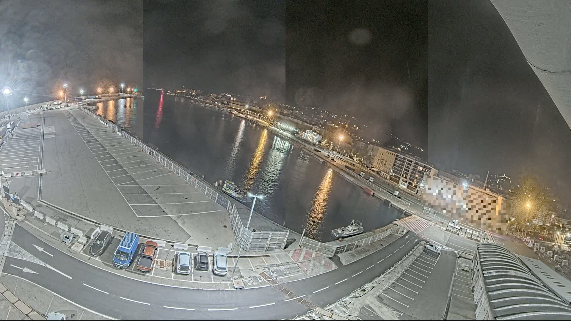 A nighttime, high-angle view of a harbor with a mostly empty parking lot, several small boats, and city lights reflecting on the water.
