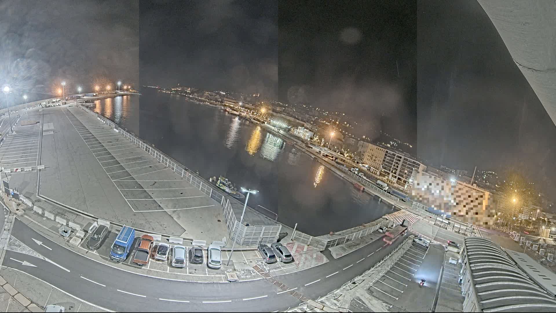A nighttime, high-angle view of a harbor with a mostly empty parking lot, a few cars, and a body of water reflecting city lights.