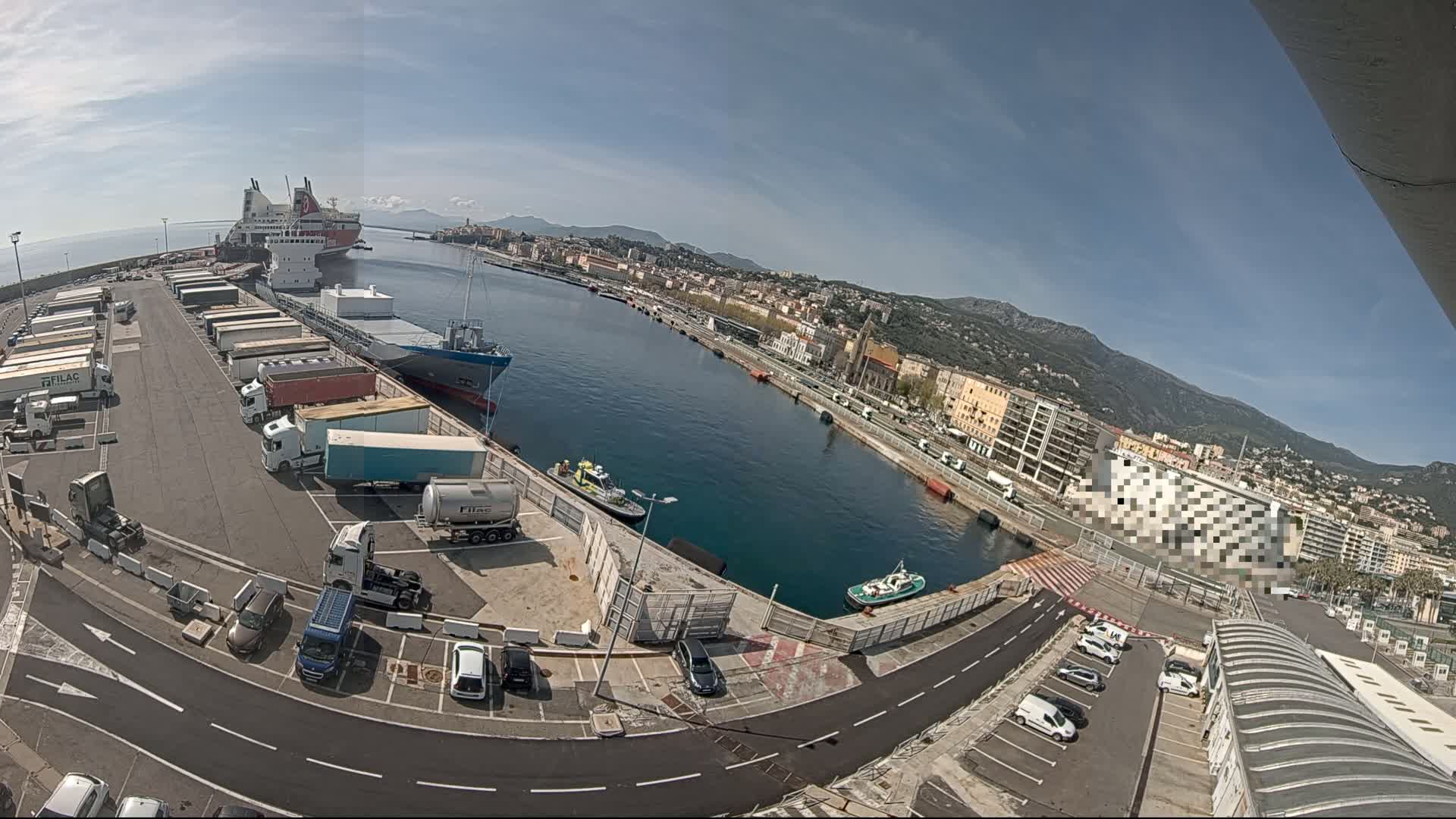 A sunny day at a port shows several ships docked, numerous trucks and cars parked in a lot, and a city backdrop with mountains in the distance.