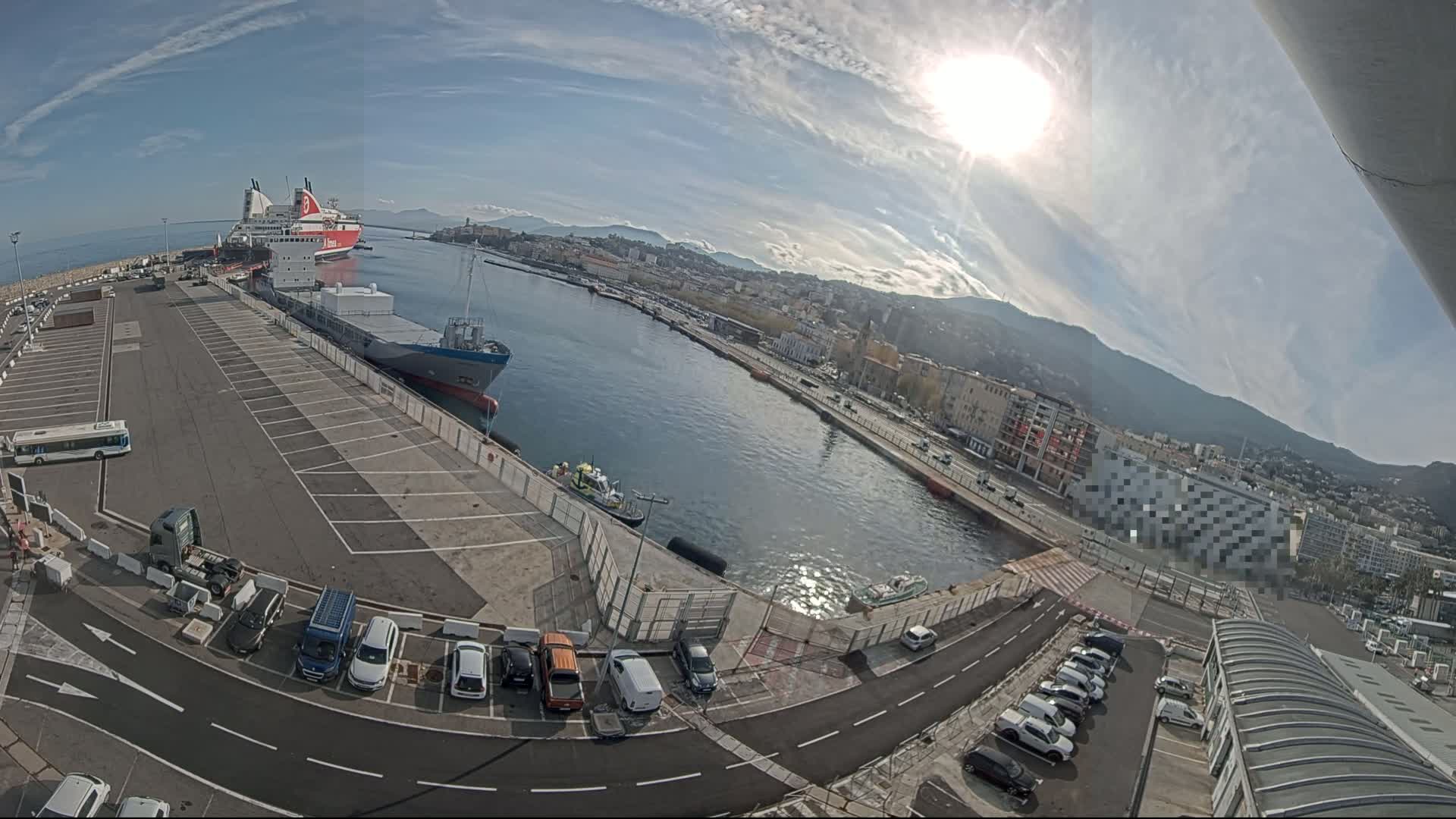 A sunny day at a harbor shows a cargo ship and ferry docked, with a parking lot and city buildings in the background.