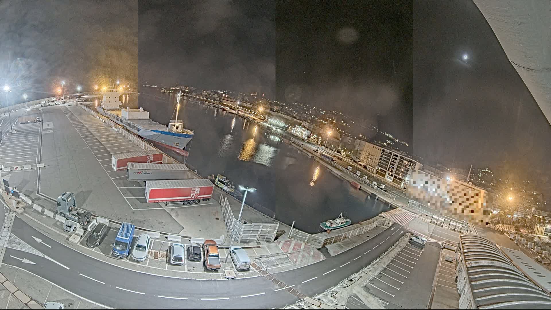 A nighttime aerial view of a harbor shows a cargo ship docked near a parking lot filled with trucks and cars, under a mostly clear sky with a bright moon.