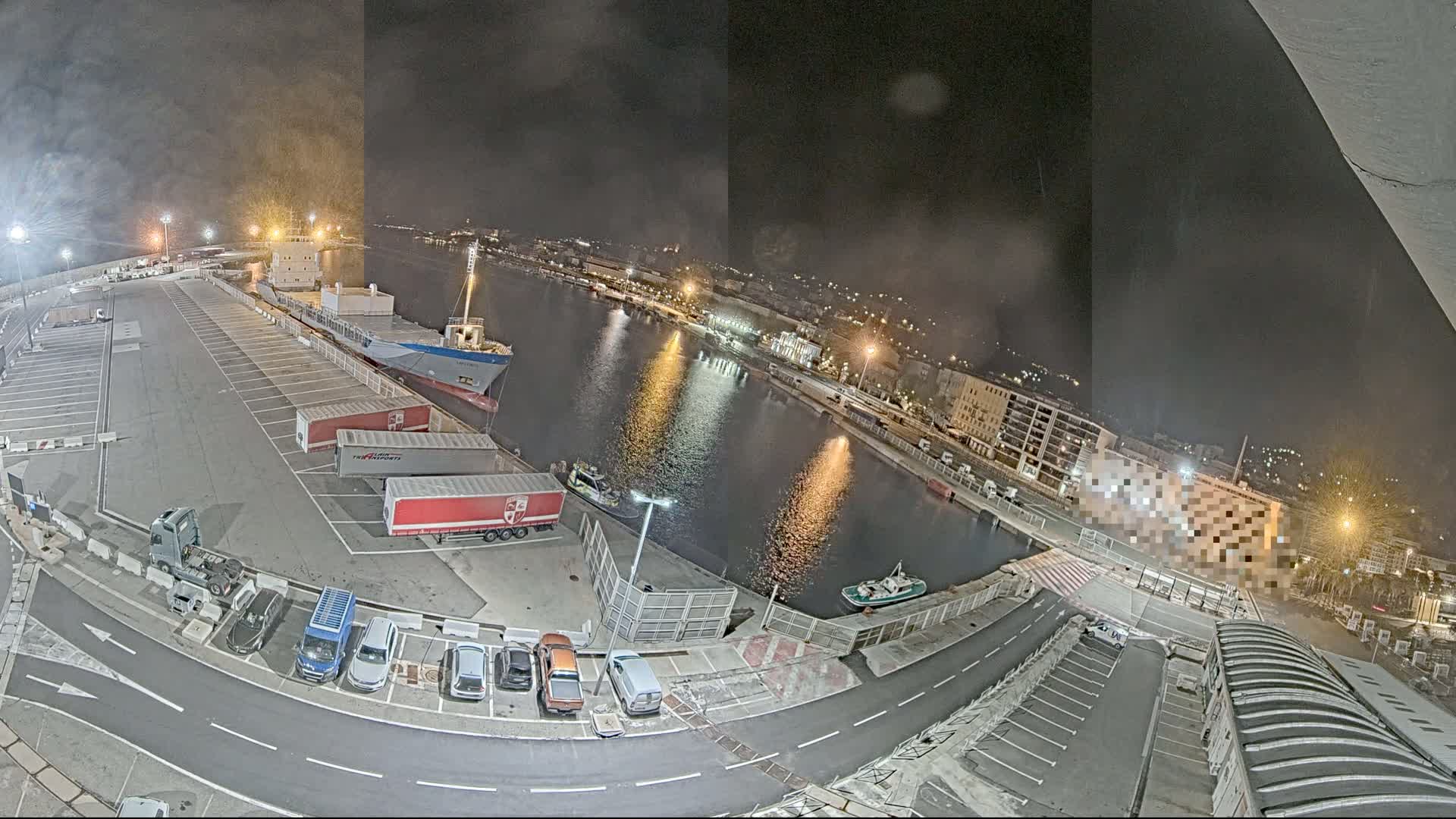 A nighttime aerial view of a harbor, showing a cargo ship docked, several trucks and cars parked in a lot, and calm water reflecting city lights.