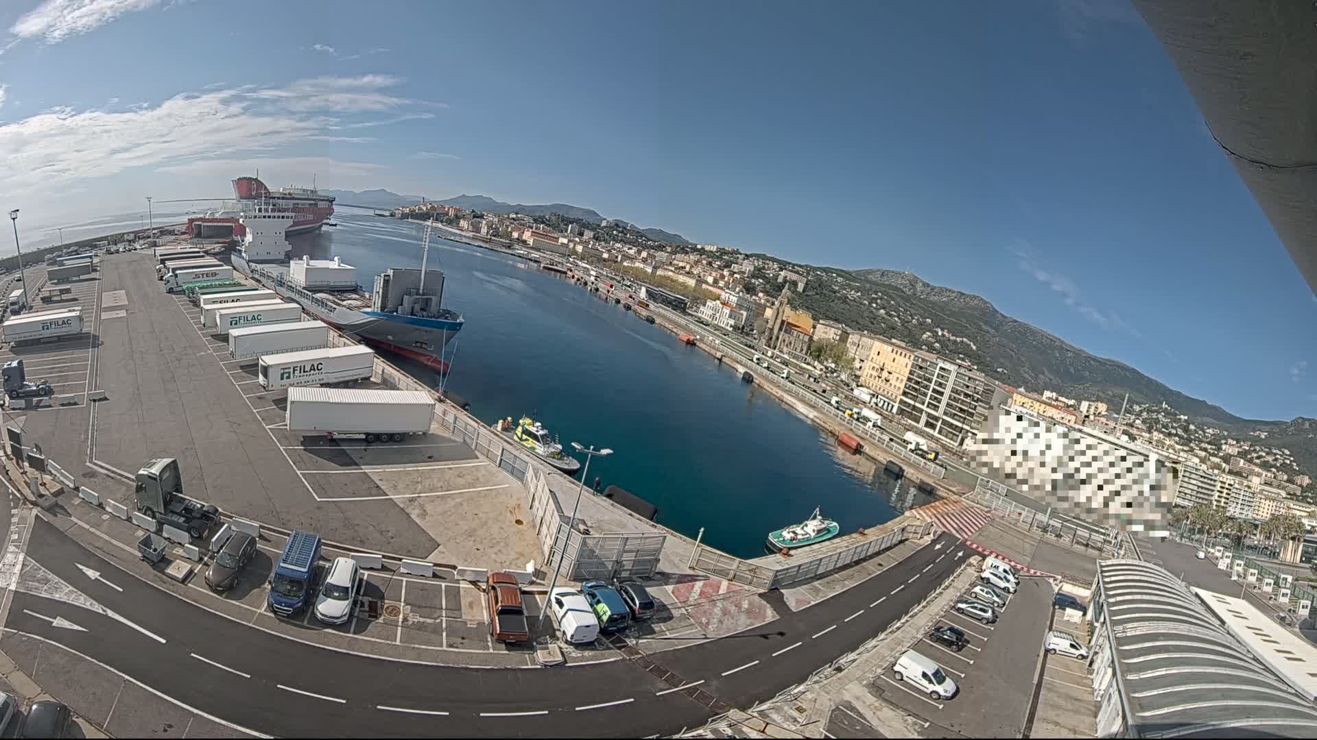 A port with several ships, numerous trucks and cars in a parking lot, and a city in the background under a clear sunny sky.