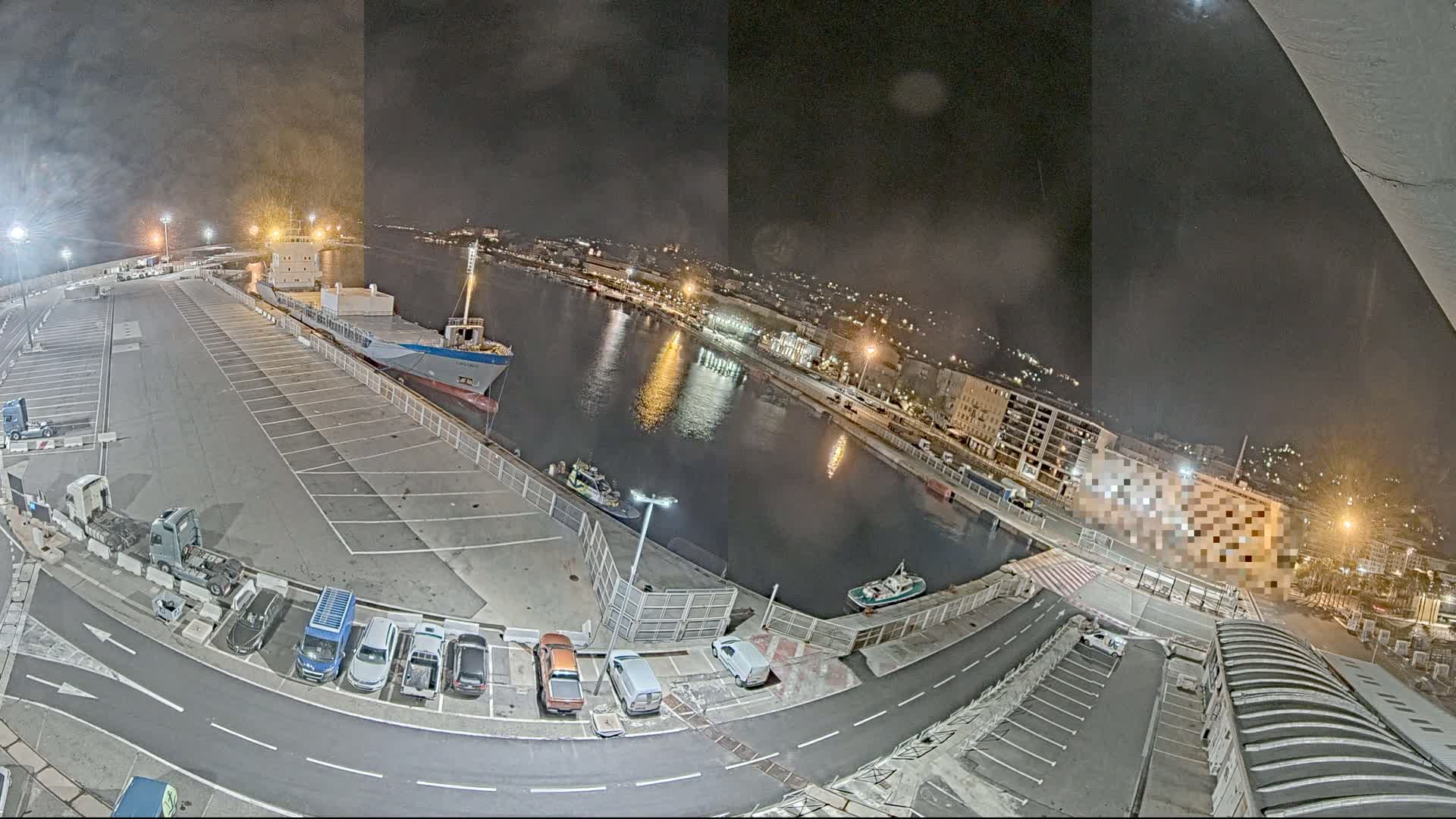 A nighttime view of a harbor with a large cargo ship docked, several smaller vessels in the water, and cars parked in a lot alongside the waterfront.