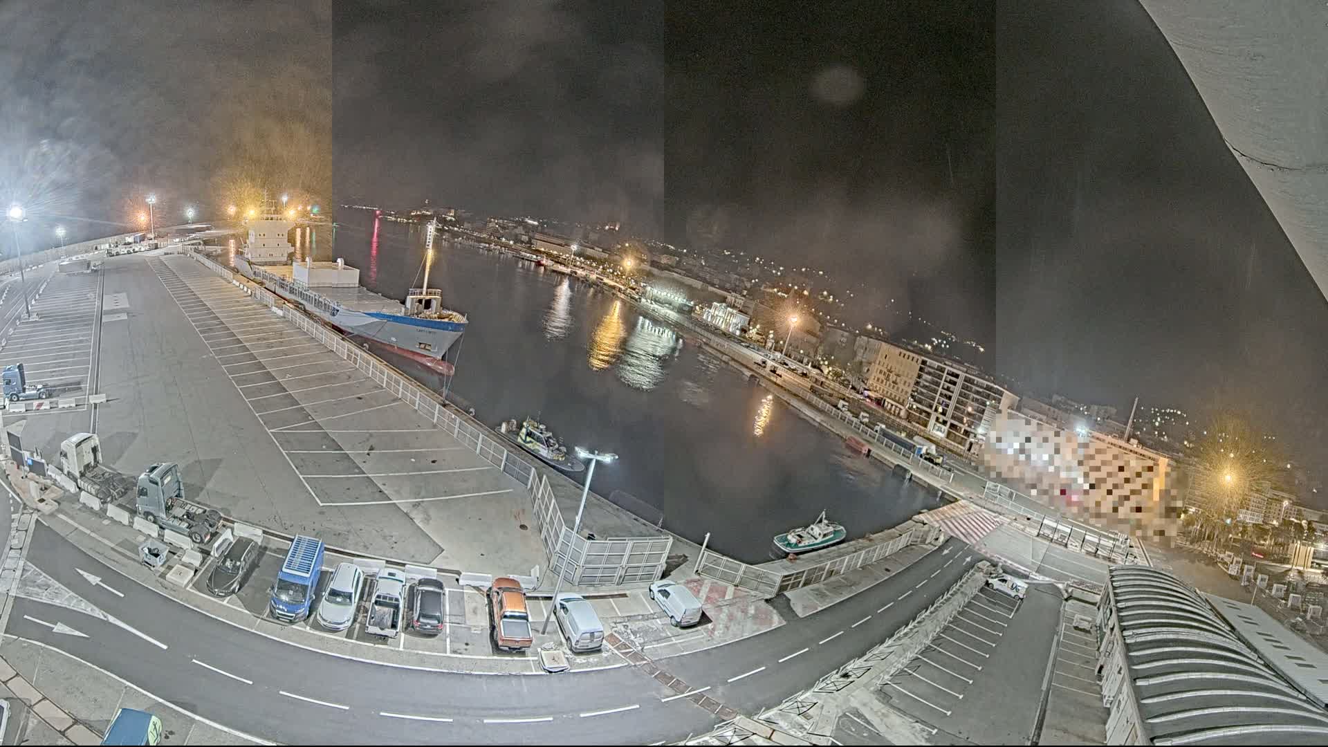A nighttime, high-angle view of a harbor with a large cargo ship docked, several smaller boats, a parking lot filled with cars and trucks, and city lights in the background.