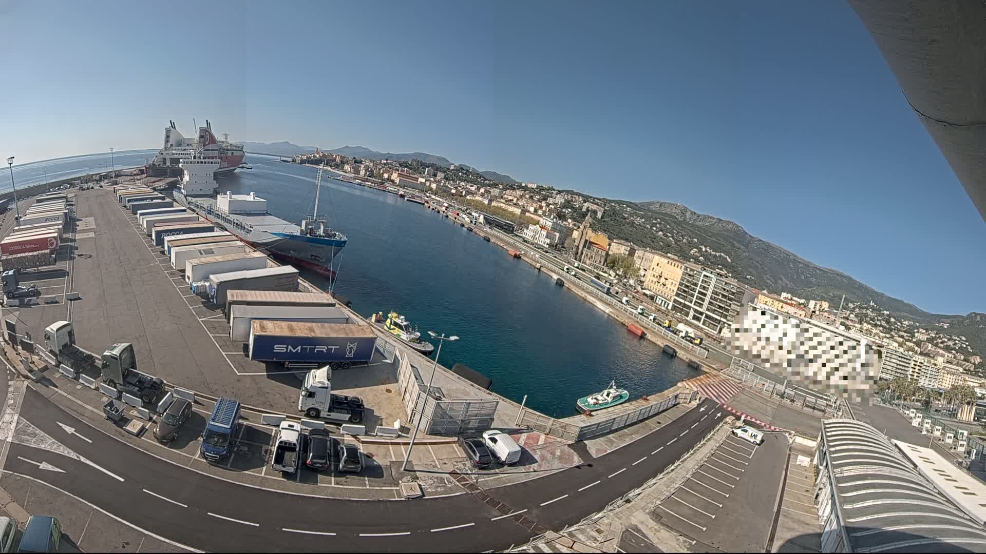 A sunny harbor scene shows several cargo ships and trailers parked at a dock next to a city nestled against a mountain range.