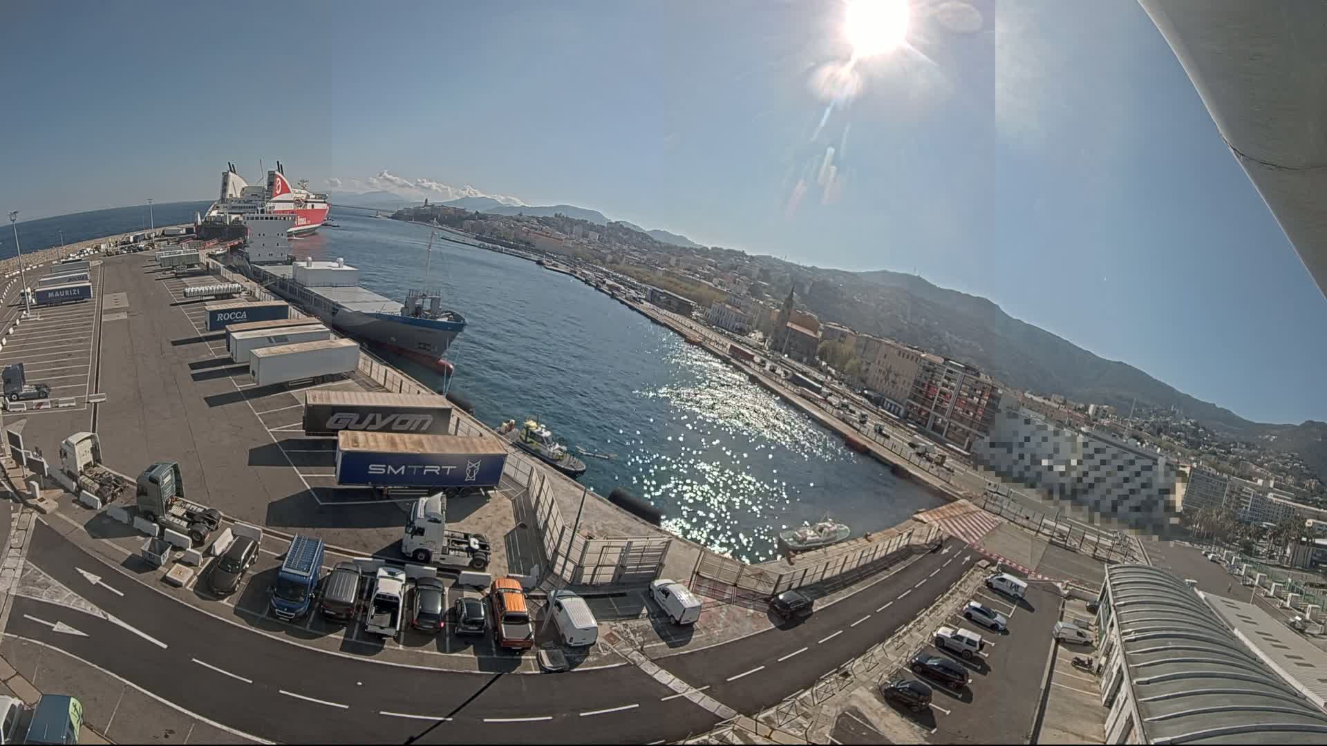 A sunny day at a harbor shows a docked ferry, cargo ships, and numerous trucks and cars parked in a lot near a city skyline and mountains in the background.