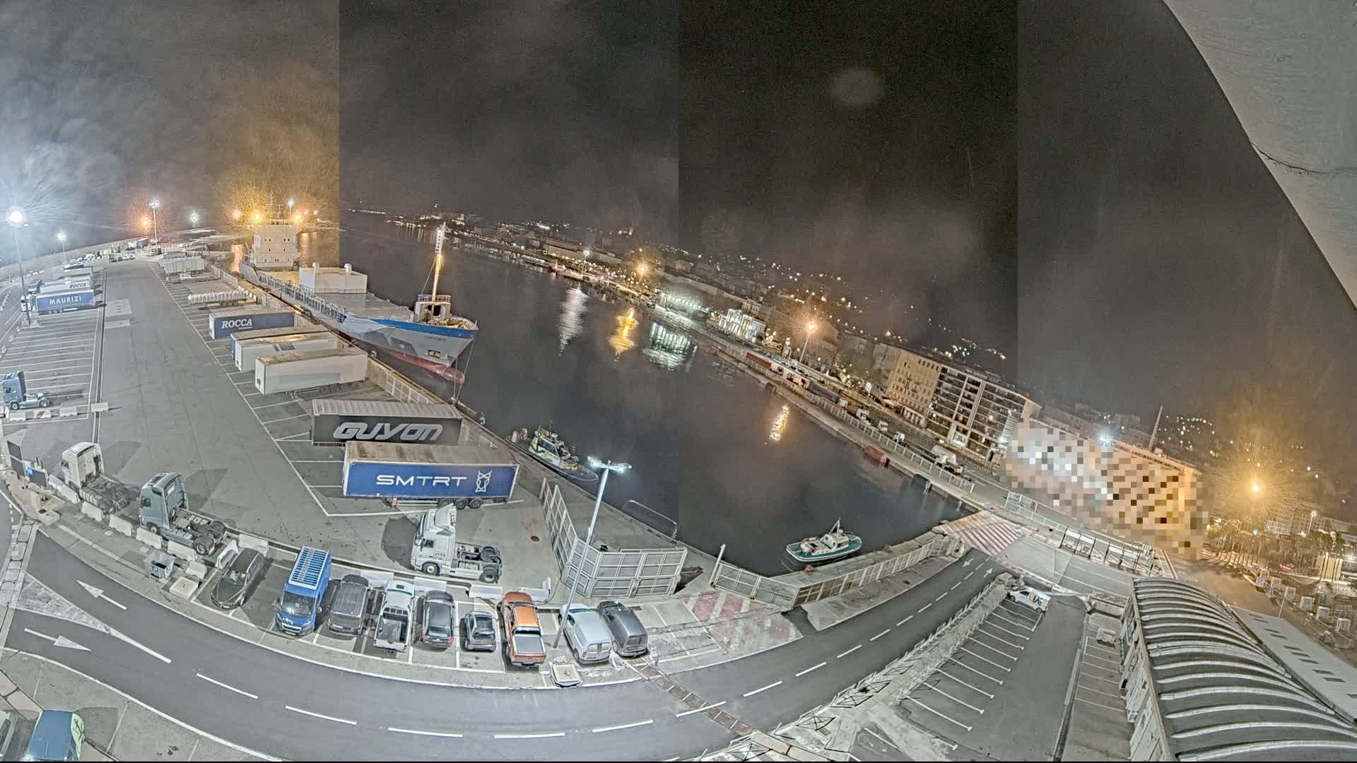 A nighttime aerial view of a harbor shows a cargo ship docked, numerous trucks and cars in a parking lot, and smaller boats in the water.