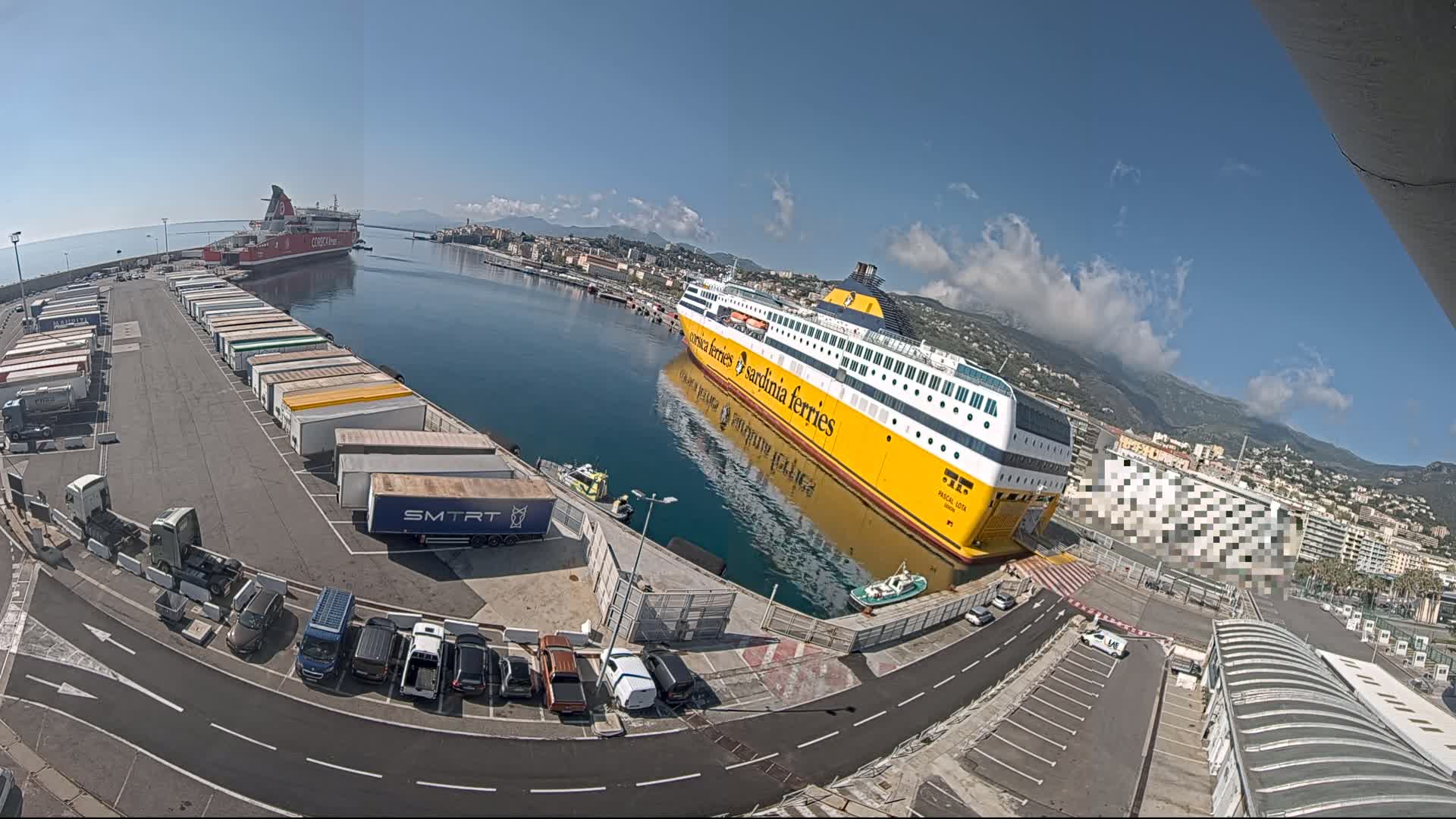 A large yellow ferry is docked at a port on a sunny day with some clouds, next to several trucks and cars in a parking lot, with another smaller ferry further down the harbor.