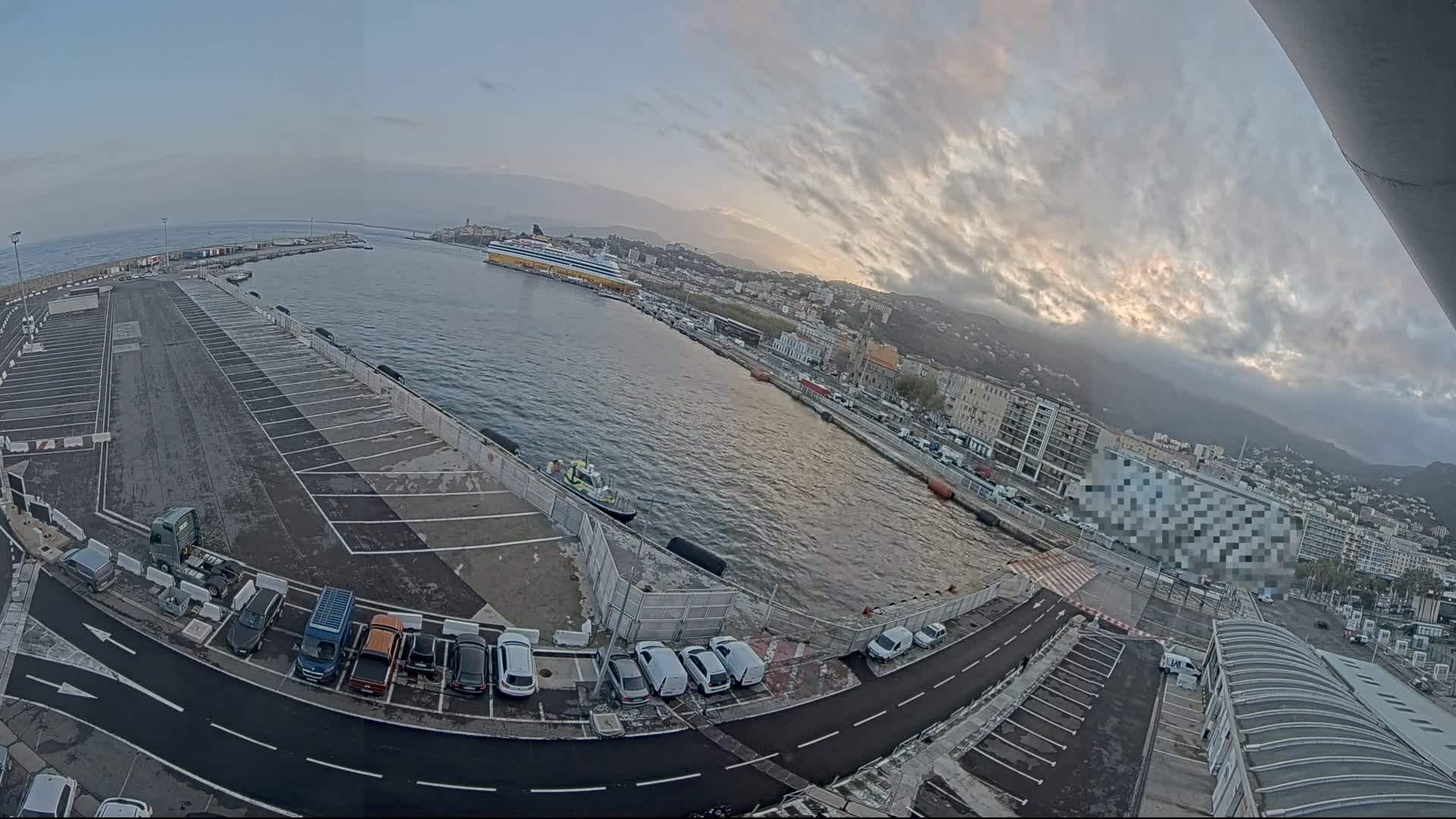 A partly cloudy sky overlooks a harbor with a ferry docked, a large parking lot with several cars, and a city in the background.