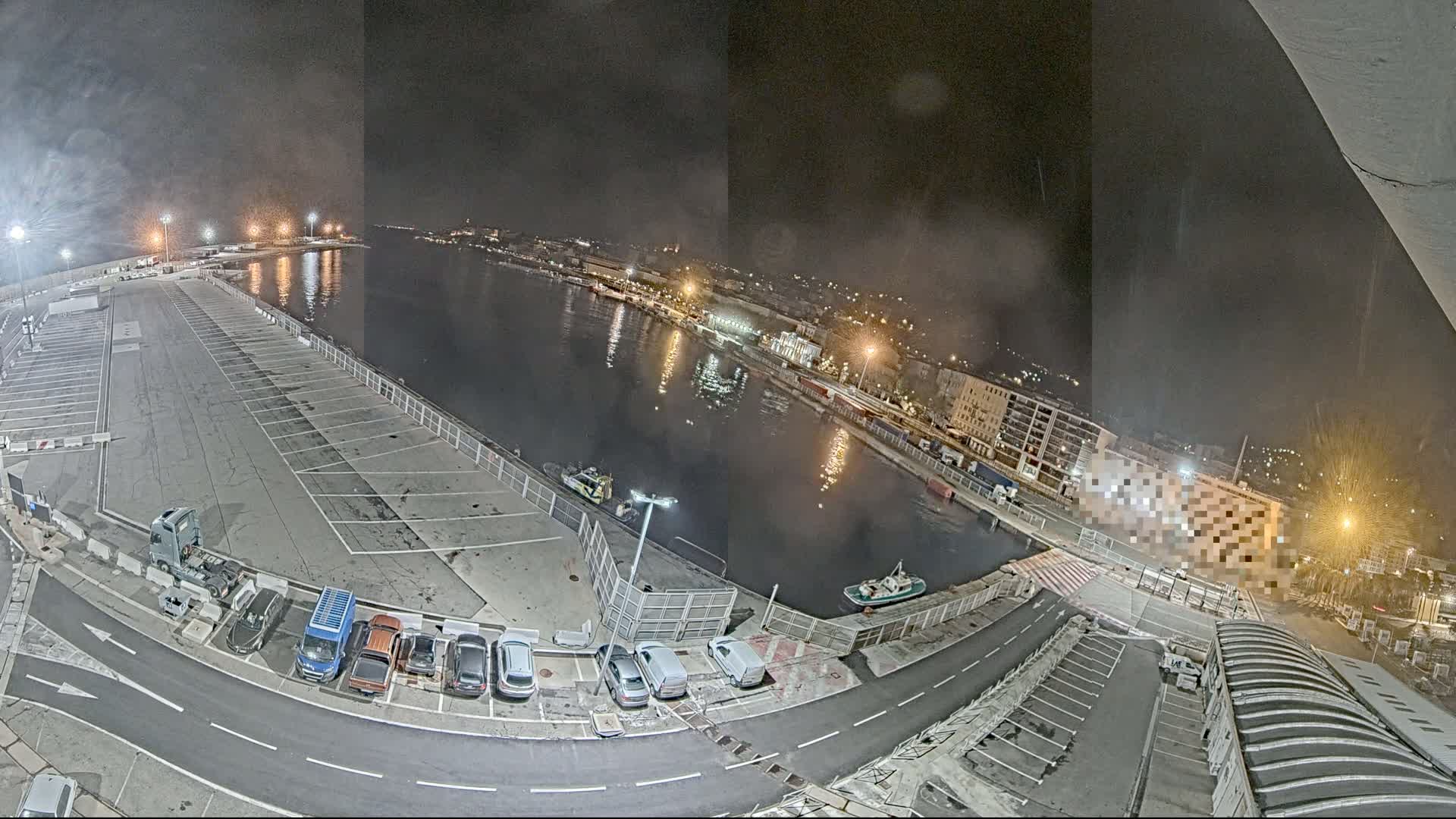 A nighttime aerial view shows a mostly empty parking lot next to a calm body of water with several small boats and a city skyline in the background.