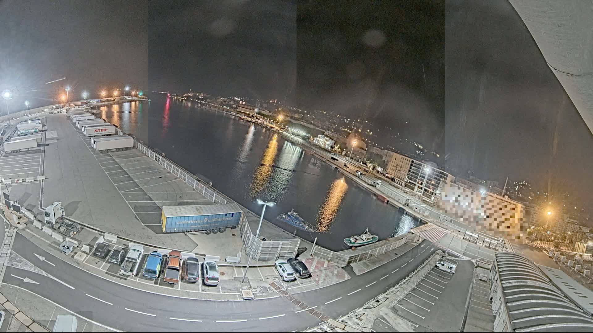 A nighttime aerial view of a harbor shows a parking lot filled with trucks and cars, a calm body of water with a few small boats, and a city illuminated by streetlights in the background.