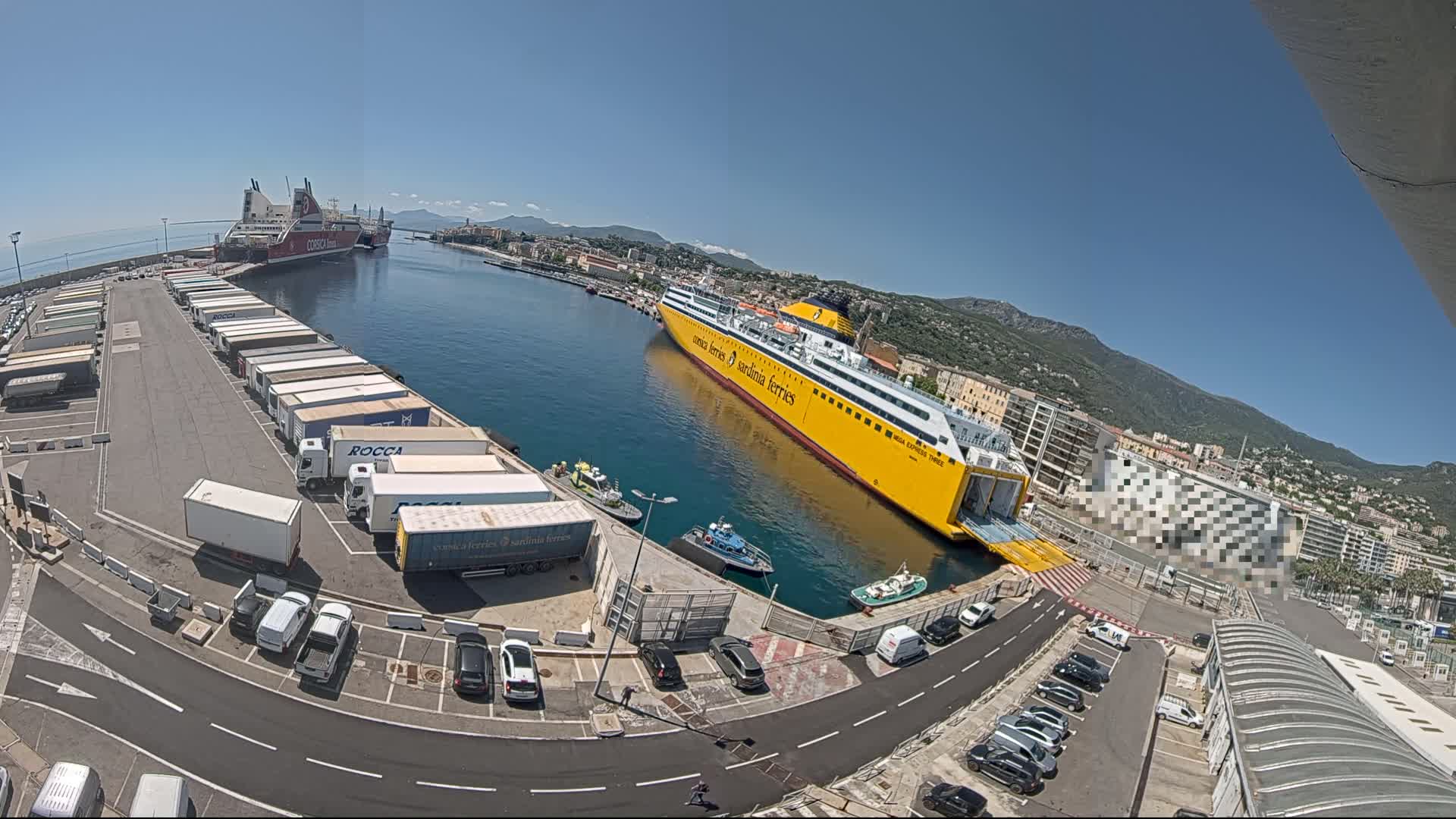 A large yellow ferry is docked at a port on a sunny day, with several other ships and numerous trucks and cars parked nearby.